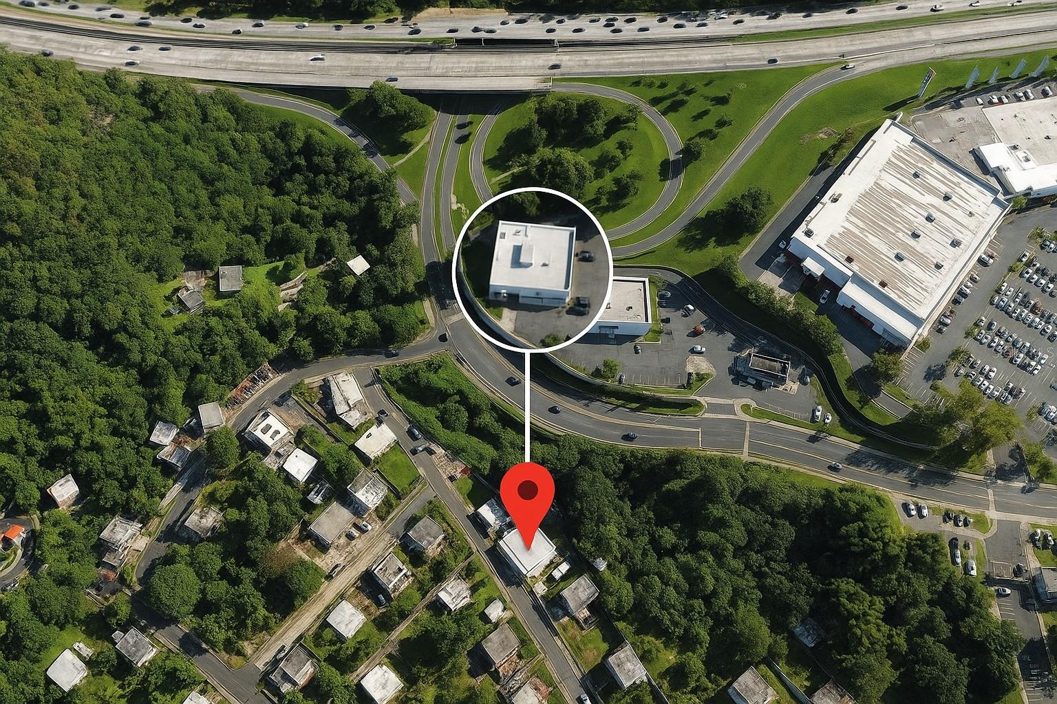 Aerial view of a small flat-roofed building marked by a red map pin amid roads and trees.