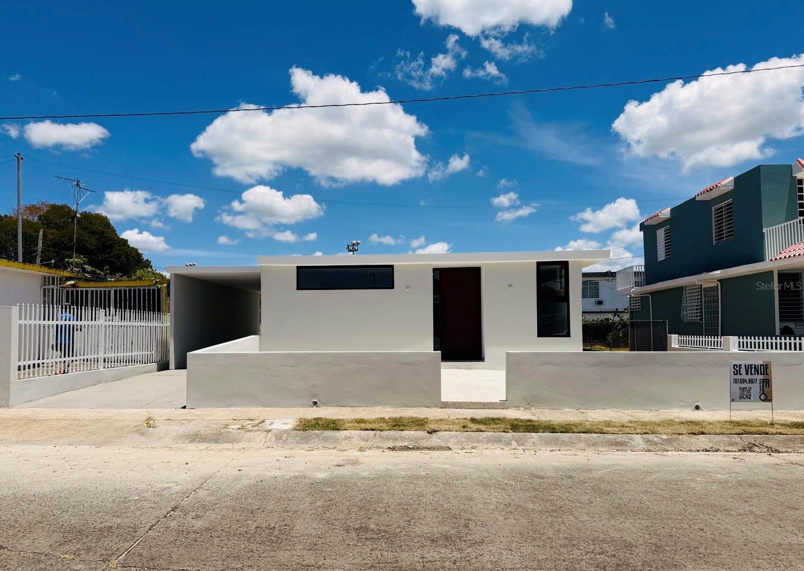 Modern white single-story house with a flat roof, covered carport, and Spanish for-sale sign.