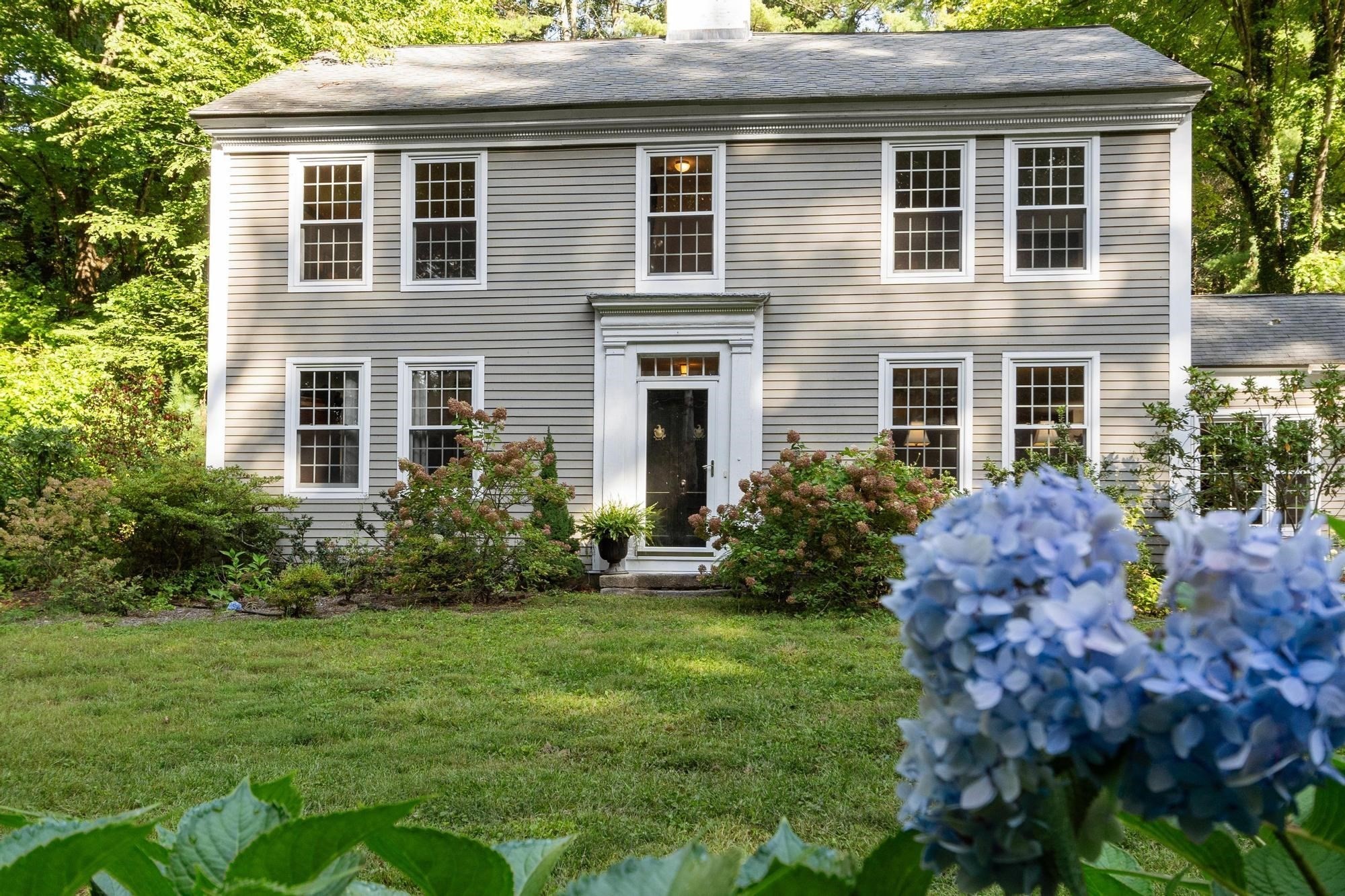 Two-story gray house with white trim, a centered black door, and blue hydrangeas in the foreground.
