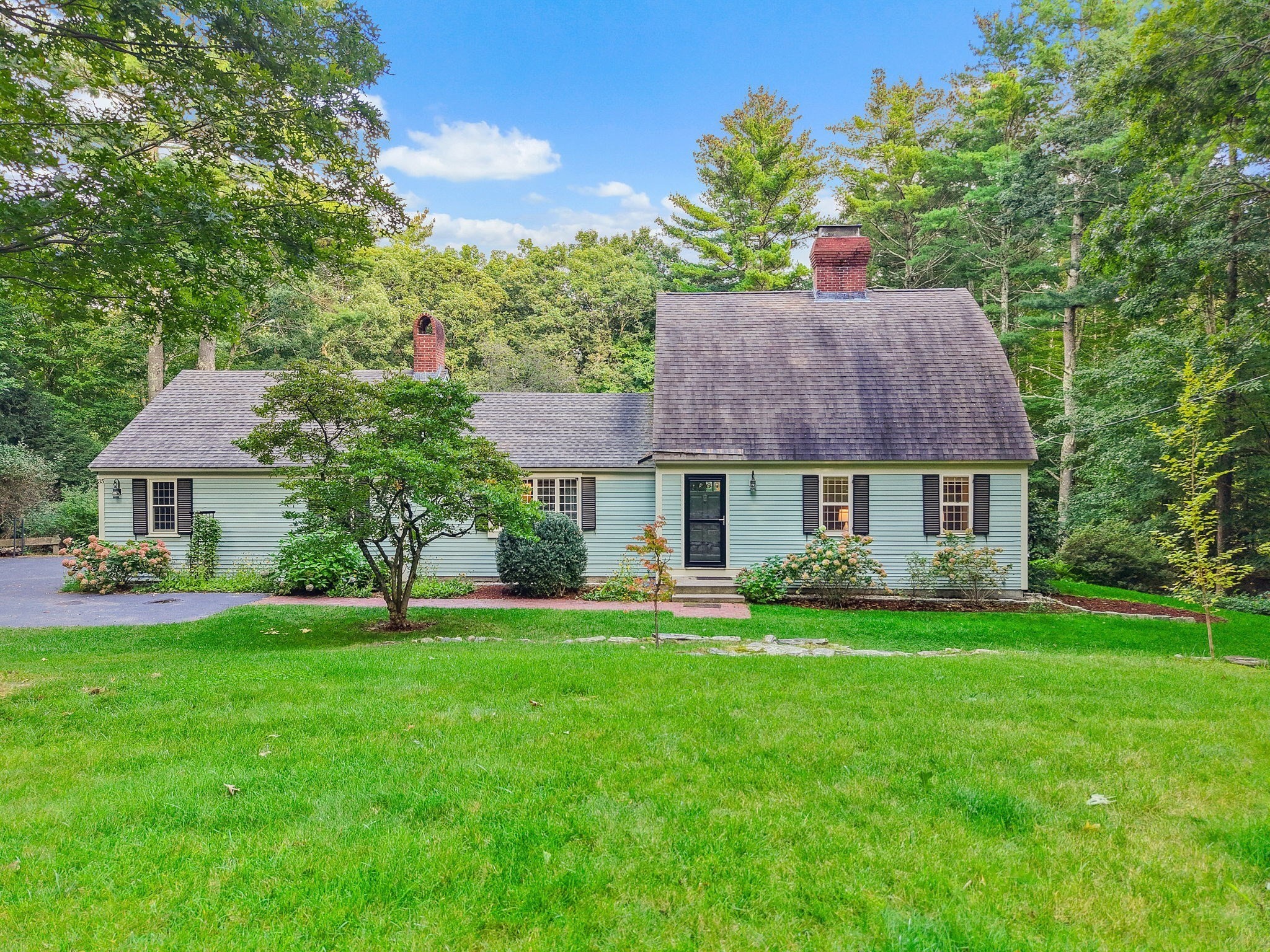 Light blue home with steep roof and brick chimneys, surrounded by mature trees and a broad green lawn.