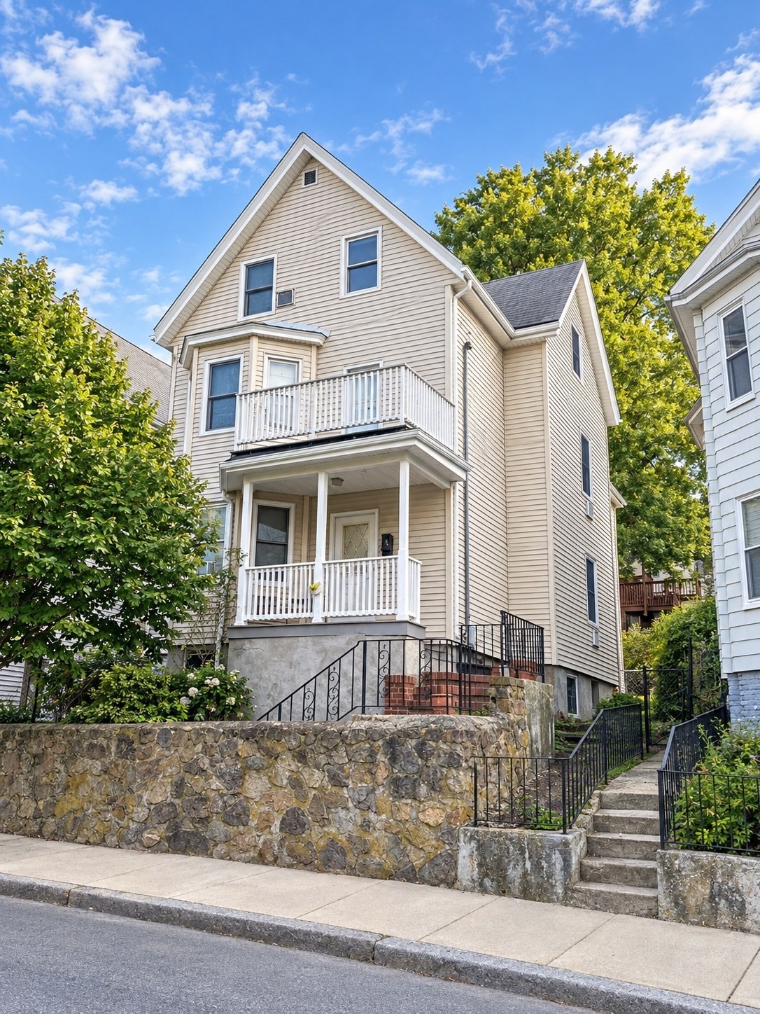 Beige multi-story house with a white balcony, covered porch, and stone retaining wall.