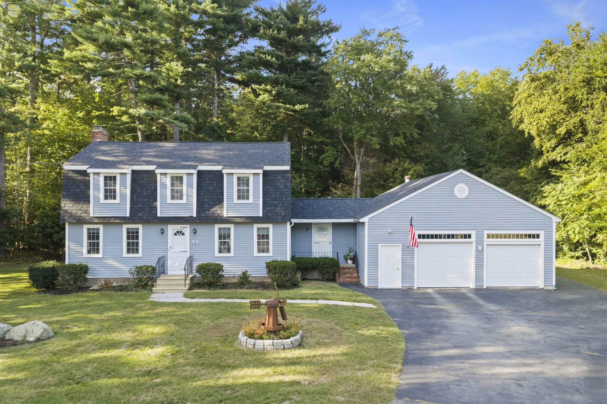 Blue-gray two-story house with dormers, an attached two-car garage, and a wooded backdrop.