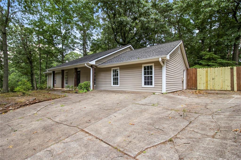 Single-story beige house with a wide concrete driveway, mature trees, and a wooden privacy fence.