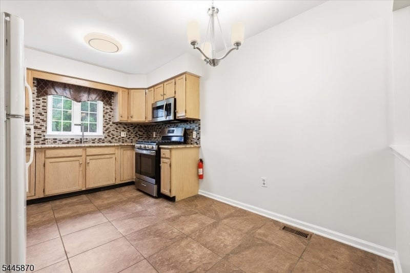 Kitchen with light wood cabinets, stainless steel appliances, and patterned tile backsplash beside an unfurnished dining area.