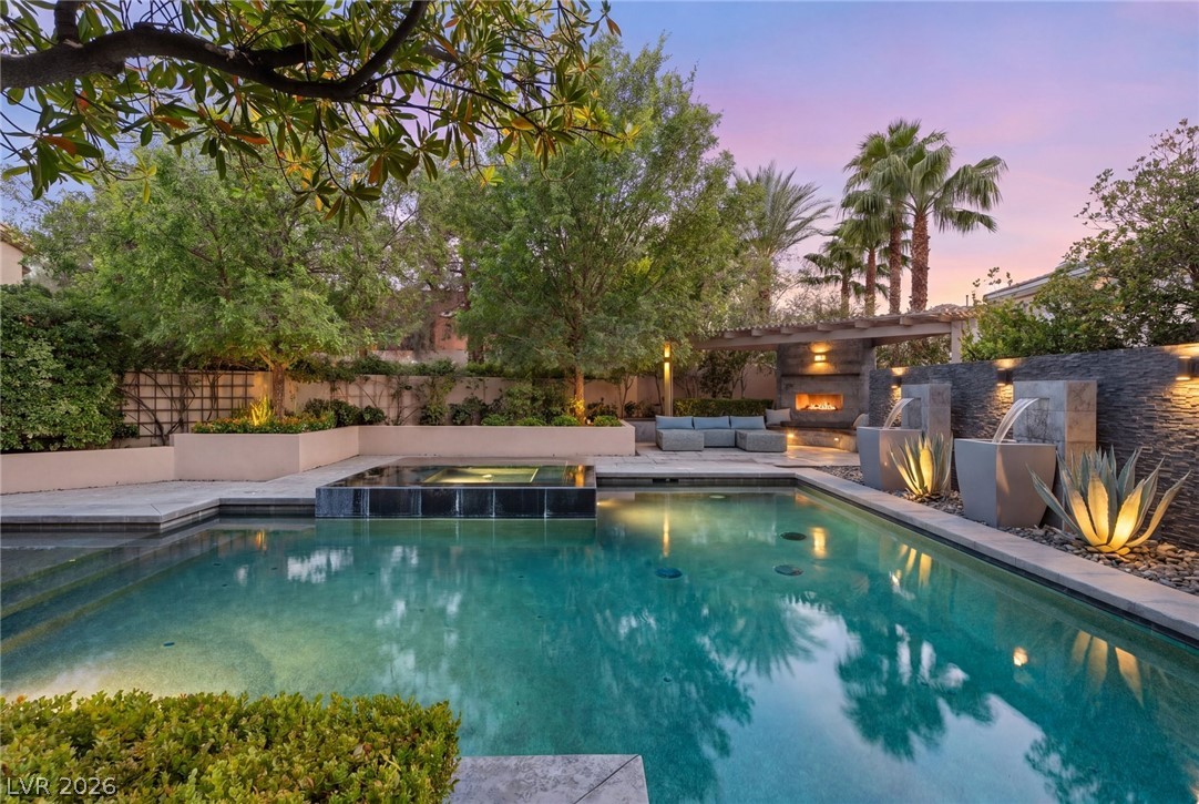 Backyard pool and raised spa beside an outdoor fireplace and built-in seating at dusk.