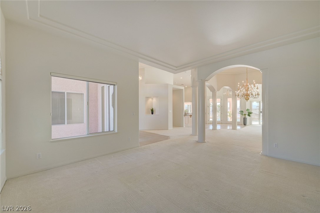 Empty living room with cream carpet, an arched opening, columns, and a chandelier.