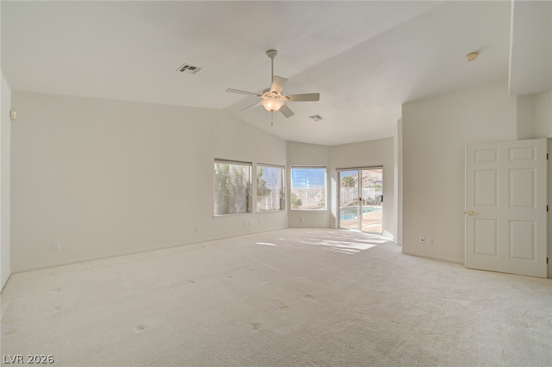 Empty carpeted living room with a ceiling fan and windows overlooking a backyard pool.