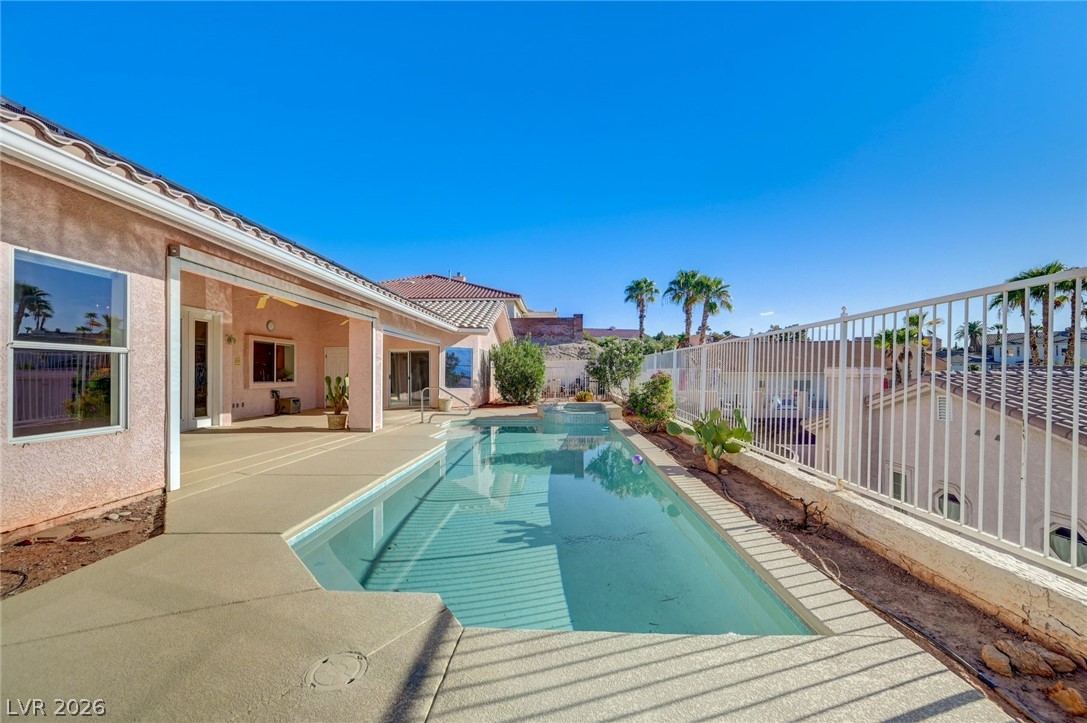 Backyard swimming pool beside a covered patio, with a white fence and palm trees beyond.