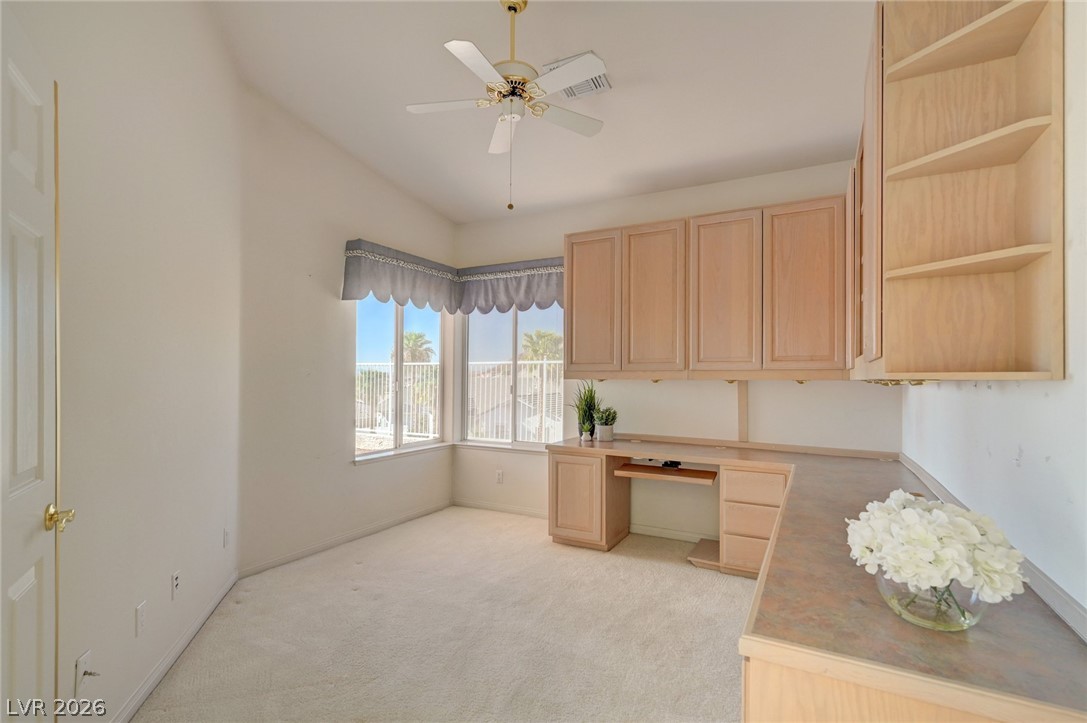 Office with built-in wood desk and cabinets, carpeted floor, and corner windows overlooking palm trees.