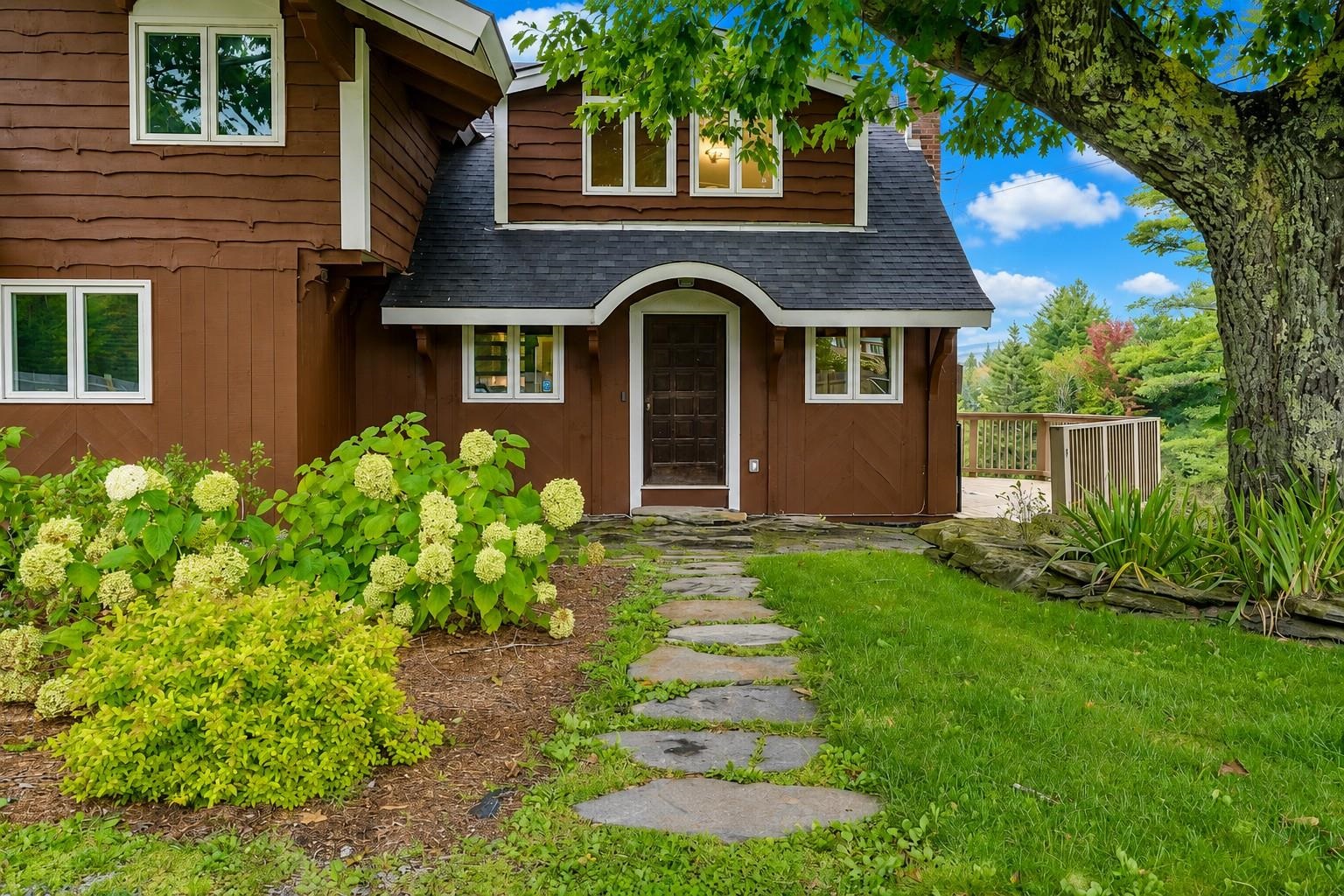 Brown shingle-sided home with an arched entry, stone path, flowering hydrangeas, and a deck.
