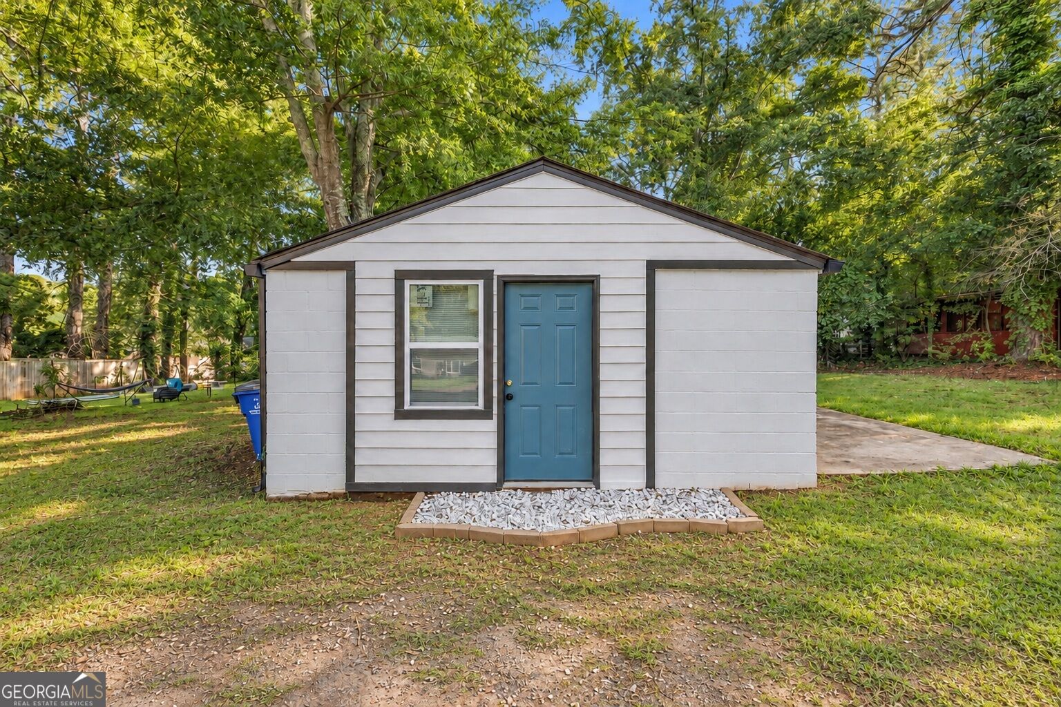 Small white-sided structure with a blue door and window, surrounded by mature trees.