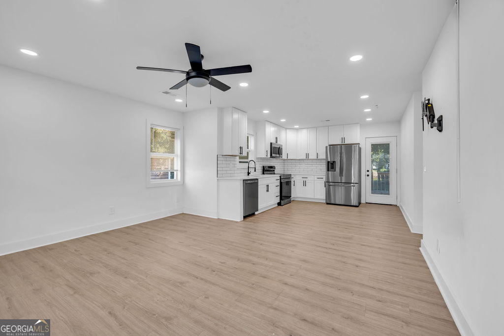 Vacant open-plan kitchen and living area with white cabinets, stainless steel appliances, and light wood flooring.