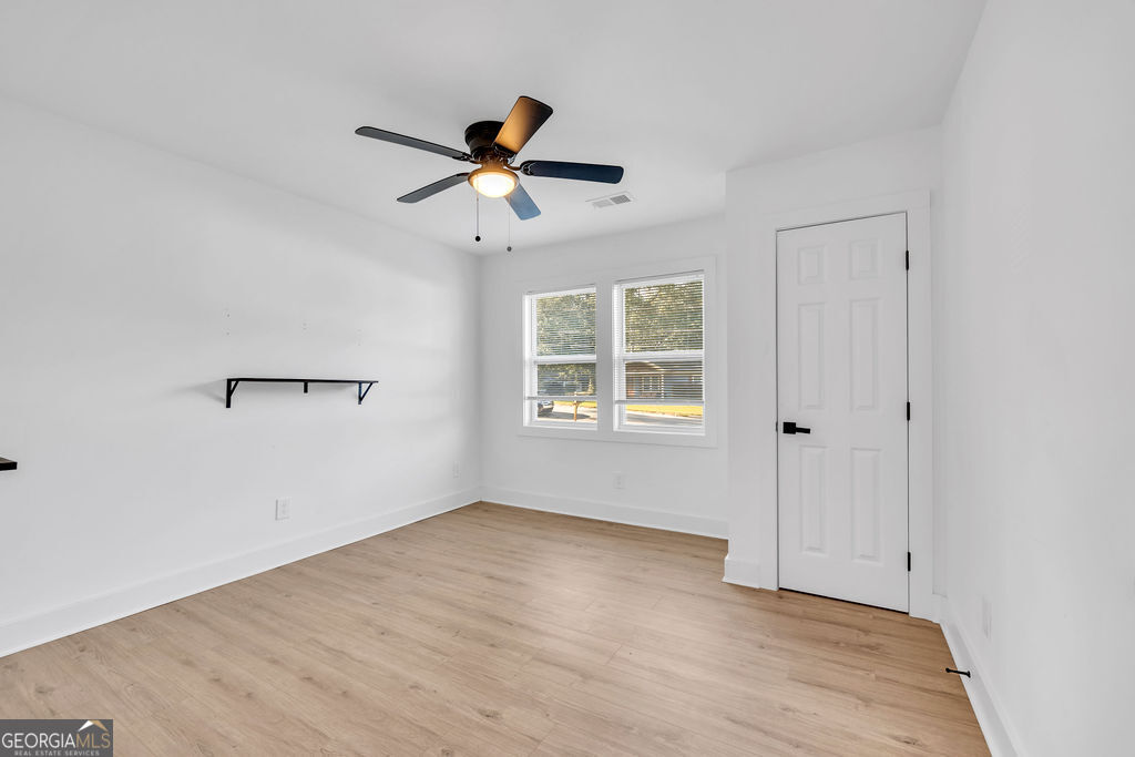Empty room with light wood flooring, a ceiling fan, two windows, and a wall-mounted shelf.