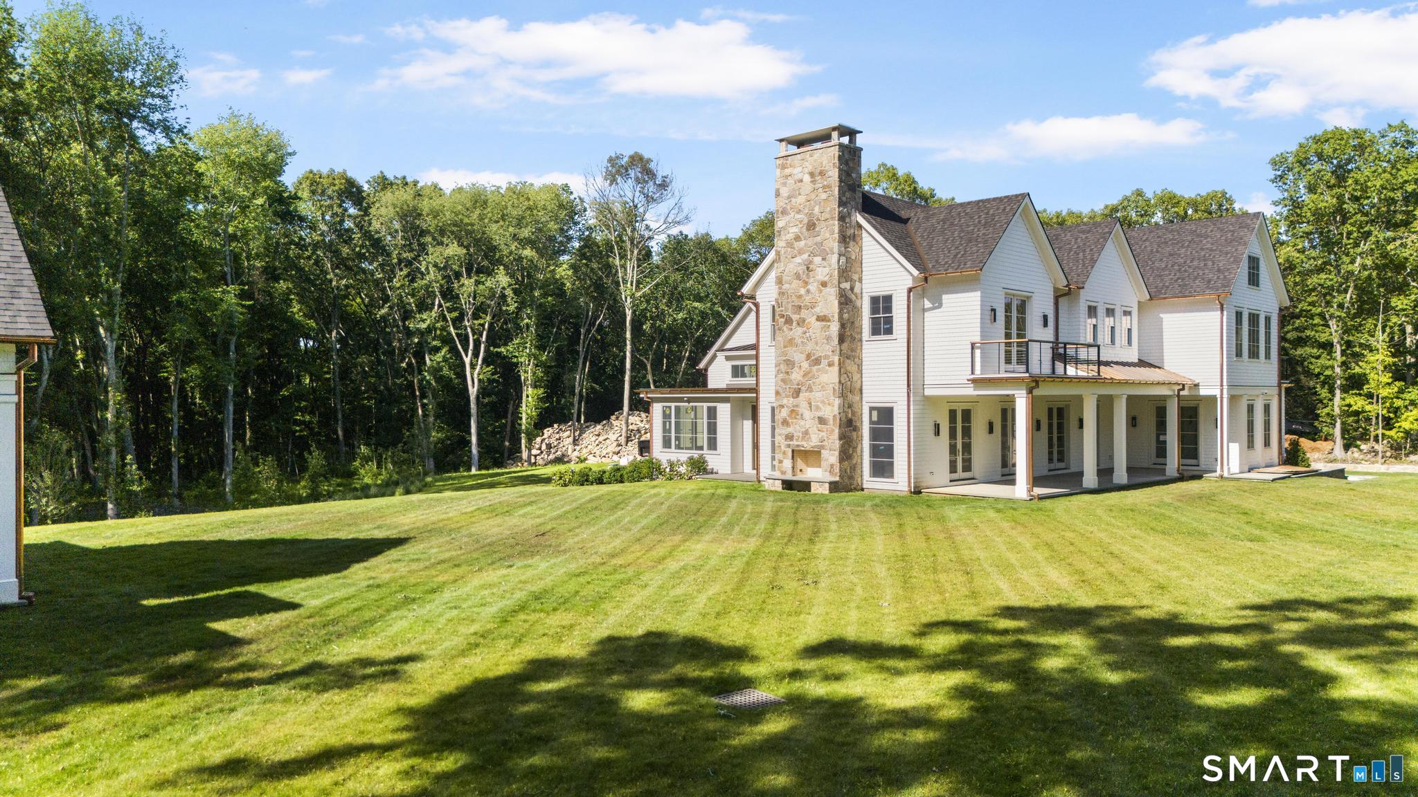 White multi-gabled home with a tall stone chimney and covered porch, set beyond a broad lawn and woods.