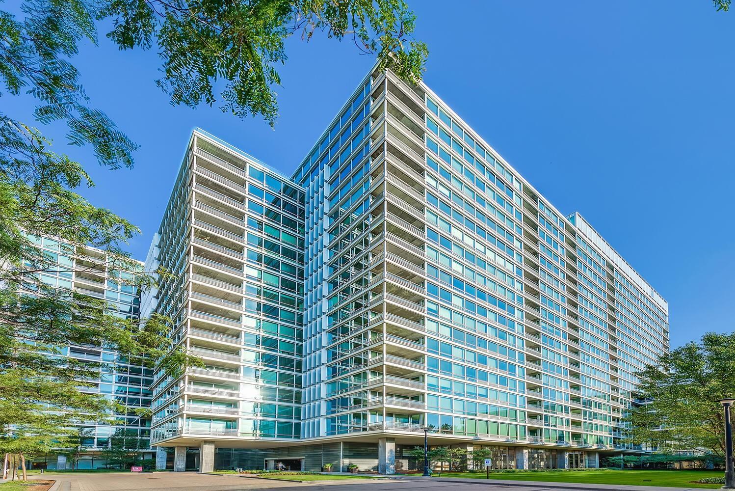 Modern glass building with angular wings, rows of windows, and trees around its landscaped grounds.