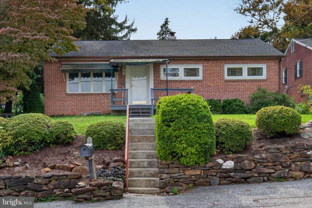 Red-brick single-story home with front steps, rounded shrubs, and a stacked-stone retaining wall.