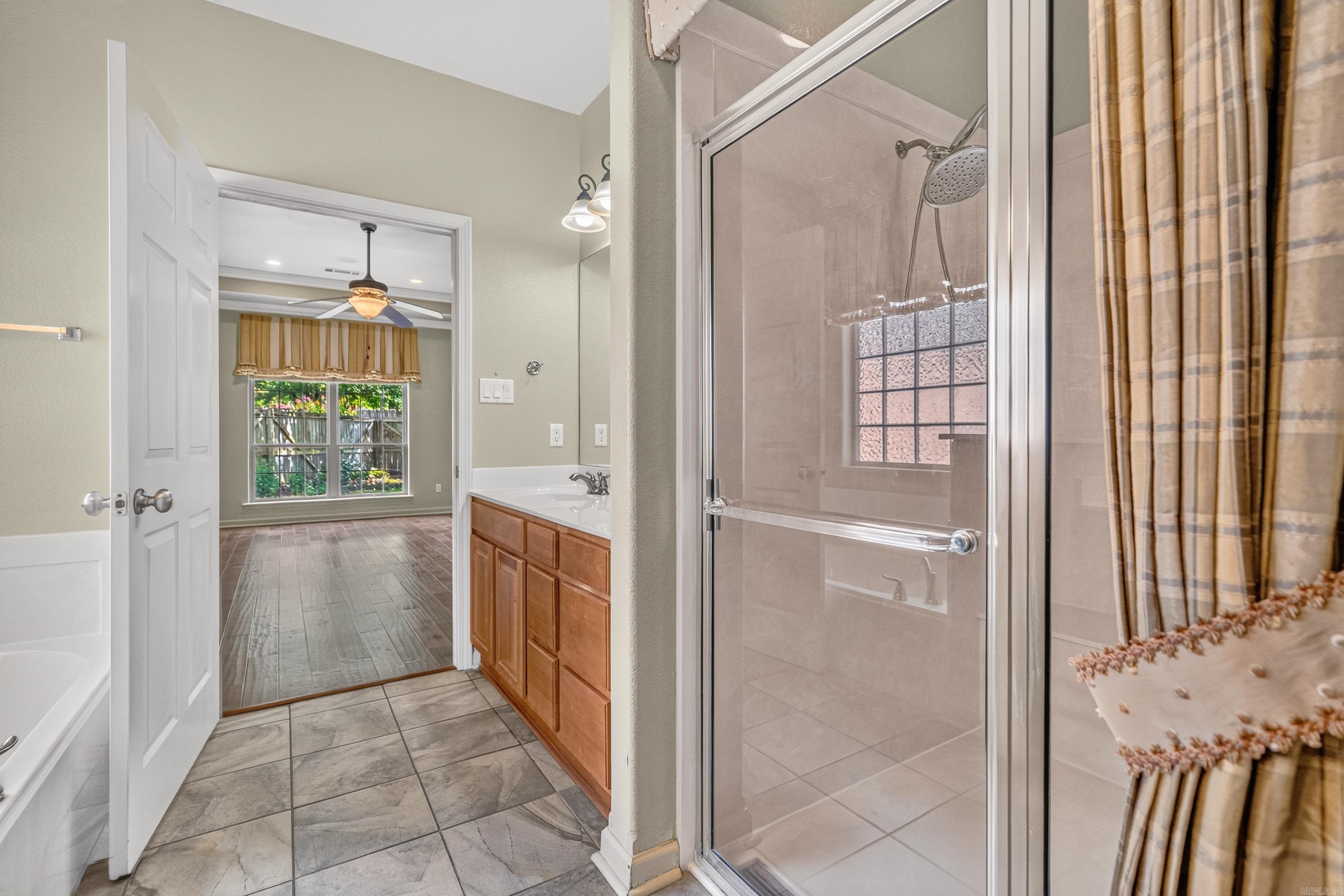 Bathroom with glass shower, bathtub, and wood vanity, opening to a room with a ceiling fan.