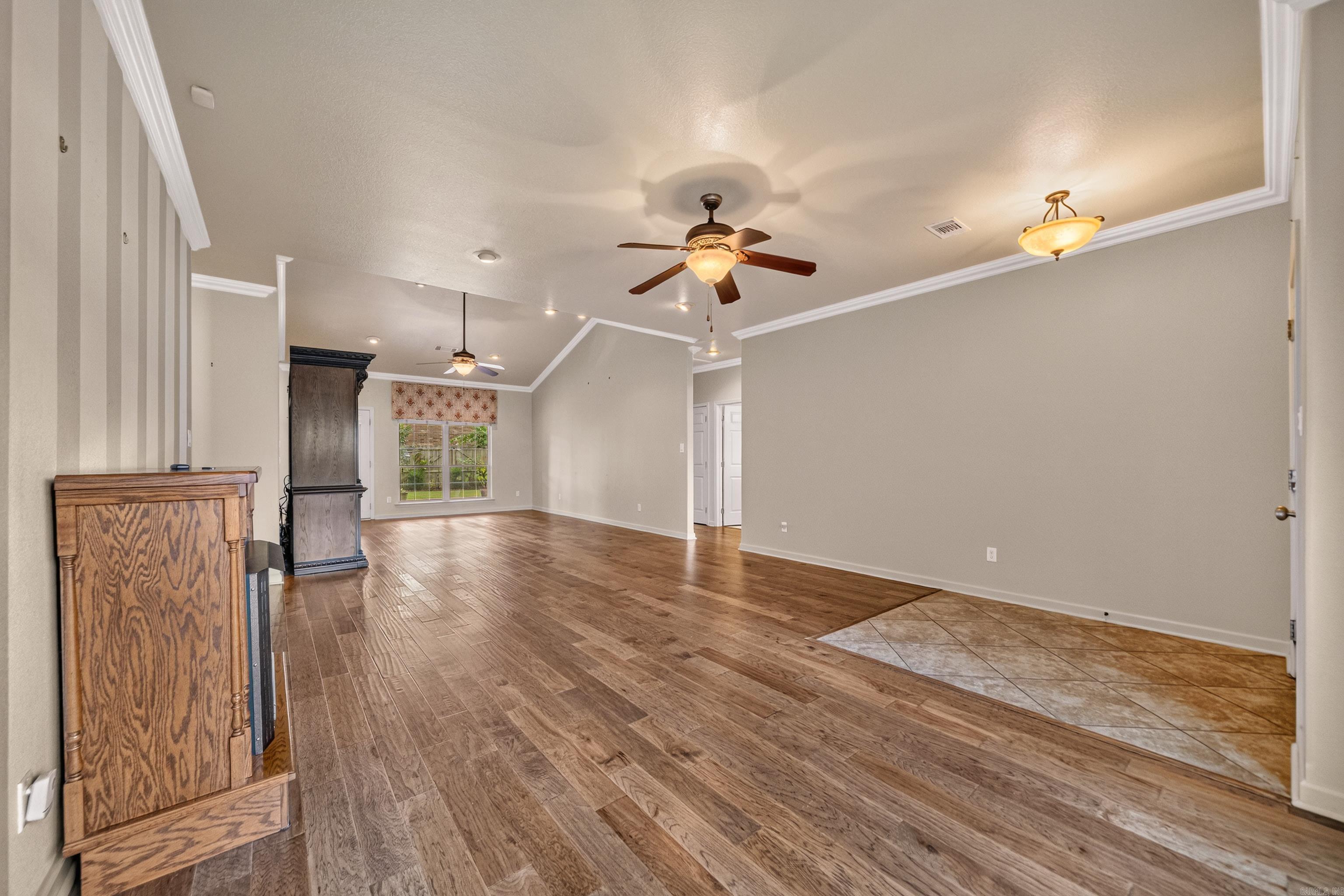 Mostly empty living room with wood-look floors, tiled entry, ceiling fans, and a window at the far end.