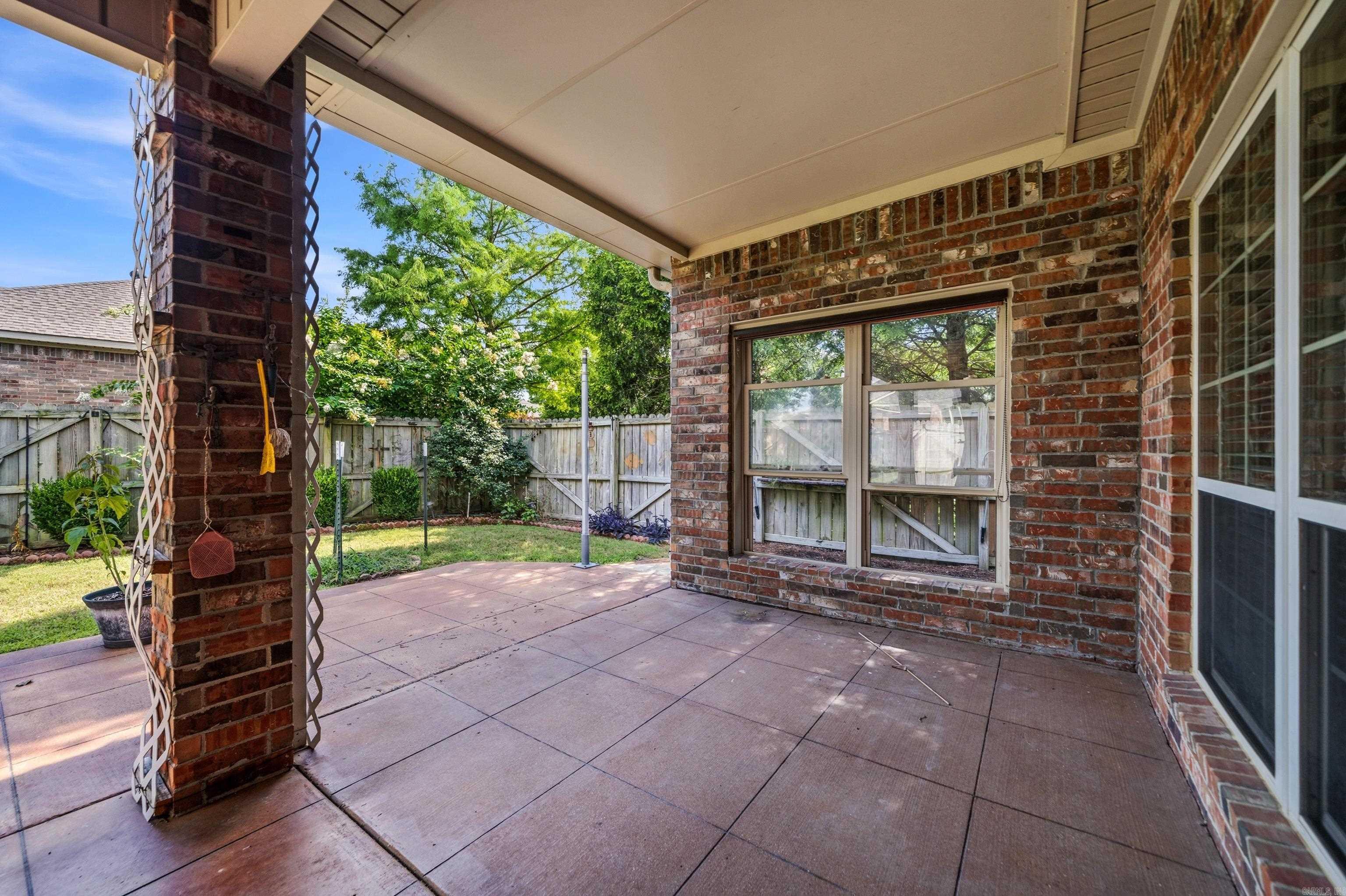 Covered concrete patio beside a brick home, overlooking a fenced backyard.