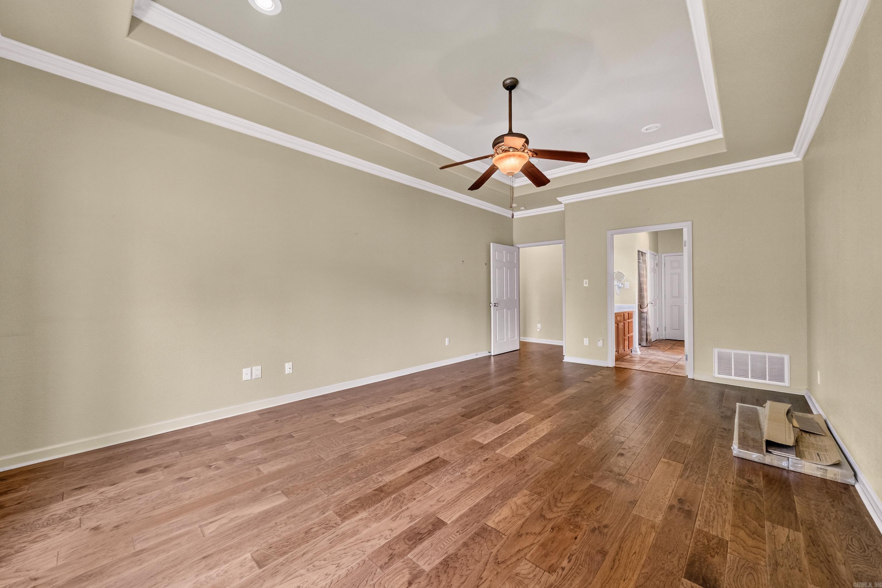 Mostly empty room with hardwood floors, a tray ceiling, and a lit ceiling fan.