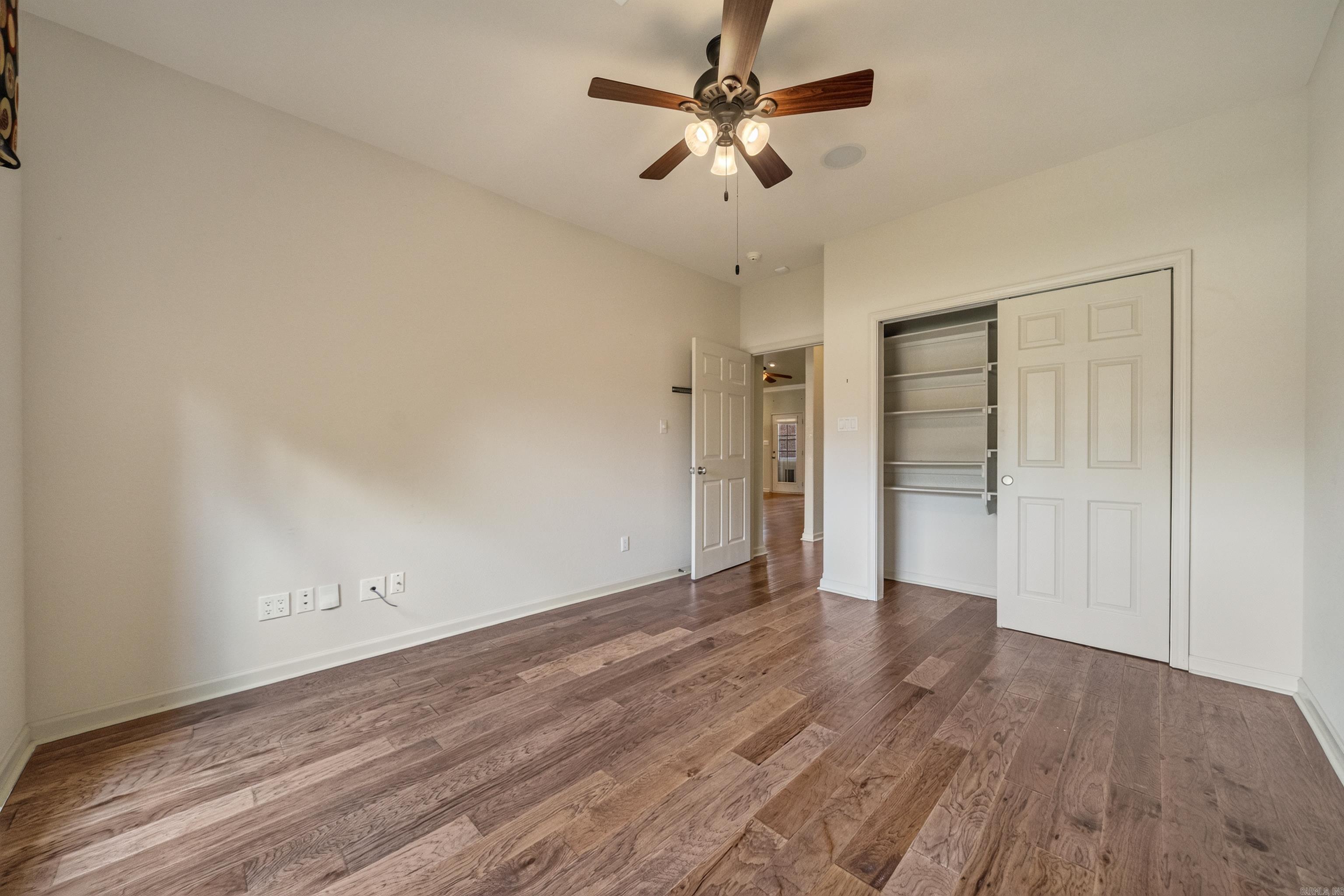 Empty bedroom with wood flooring, a ceiling fan, and an open closet with shelves.