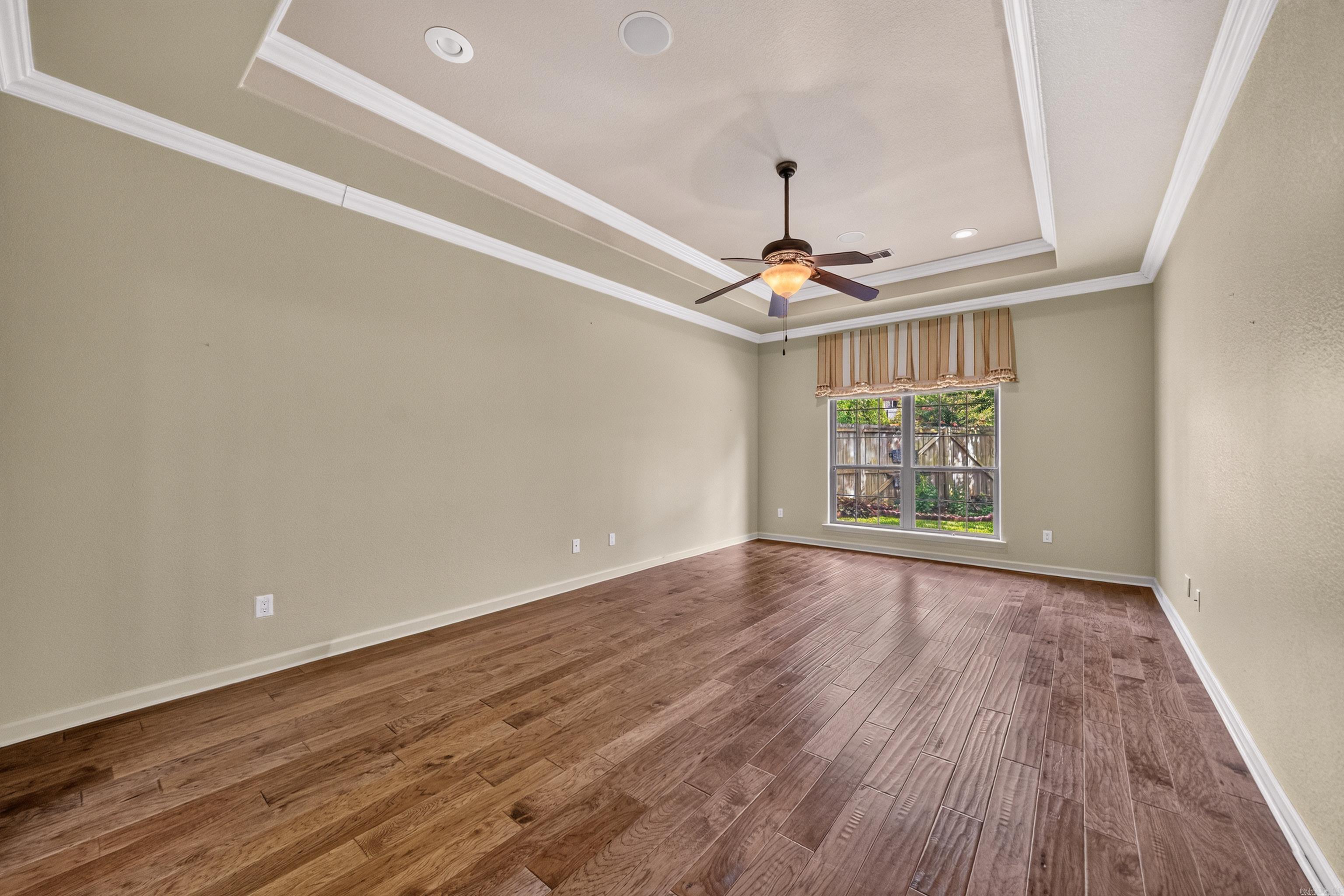 Empty room with wood plank flooring, tray ceiling, ceiling fan, and a window overlooking a fenced garden.