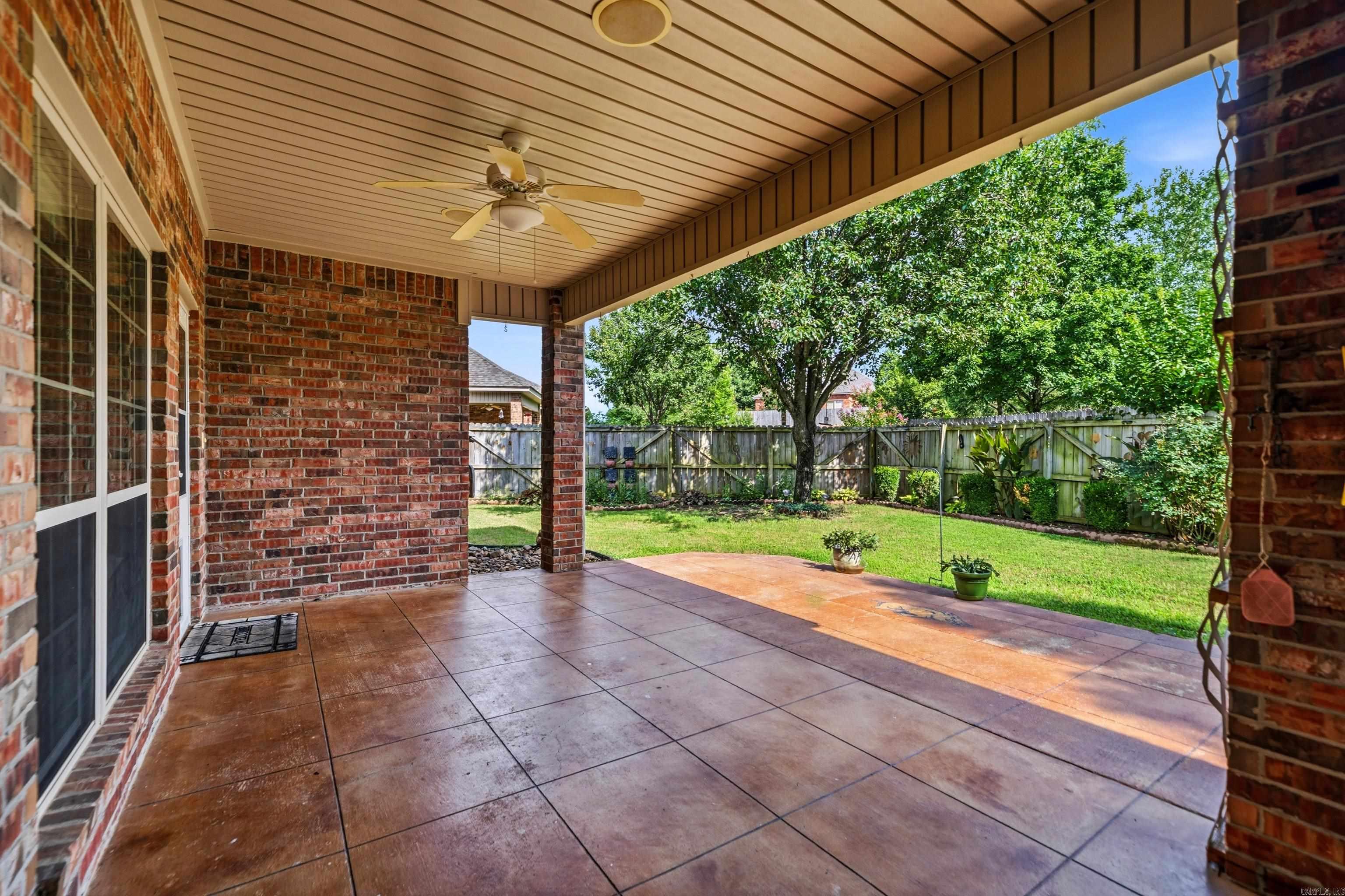 Covered brick patio with a ceiling fan overlooking a fenced, tree-shaded lawn.