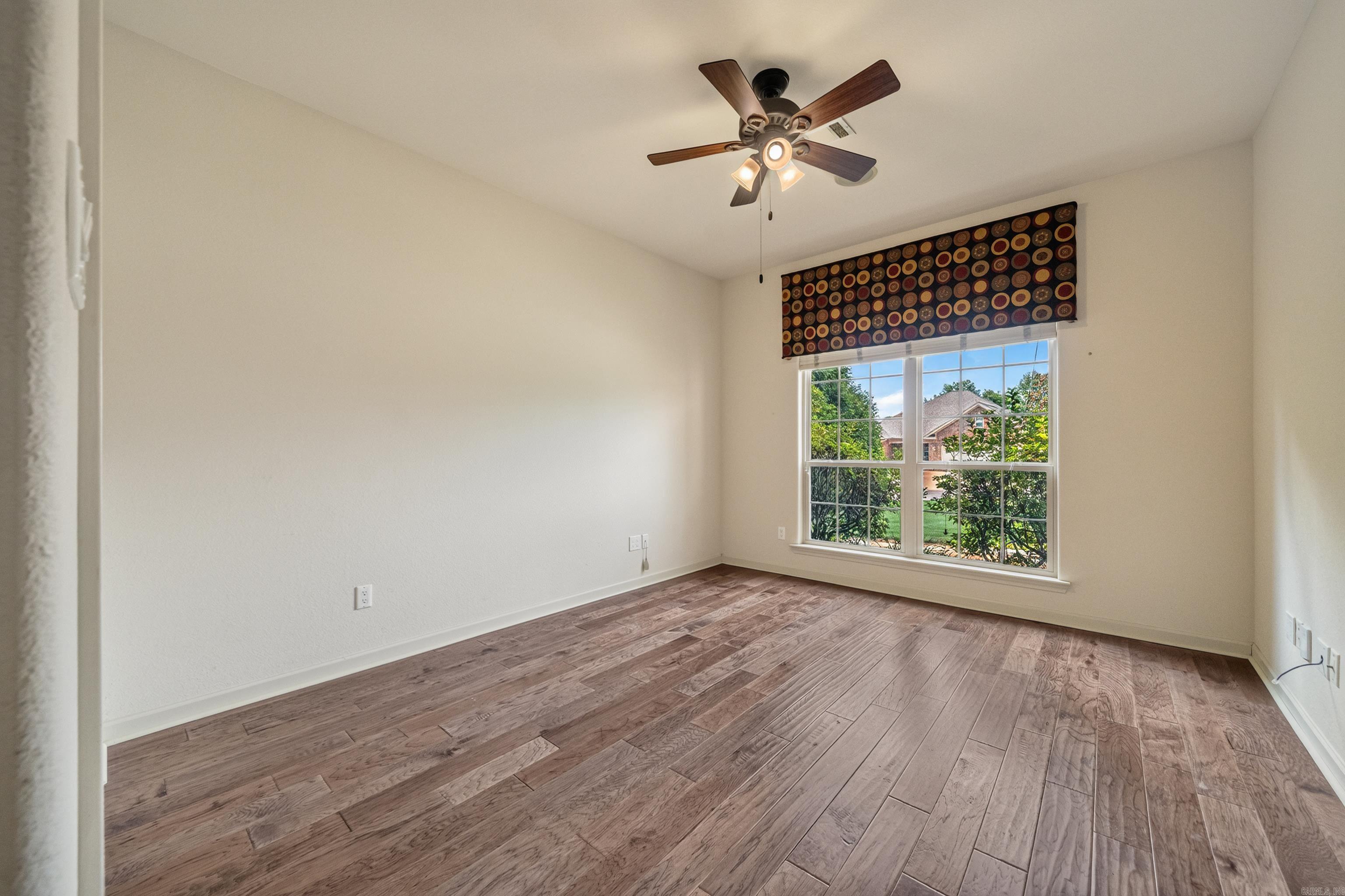 Empty bedroom with plank flooring, a ceiling fan, and a large window overlooking greenery.