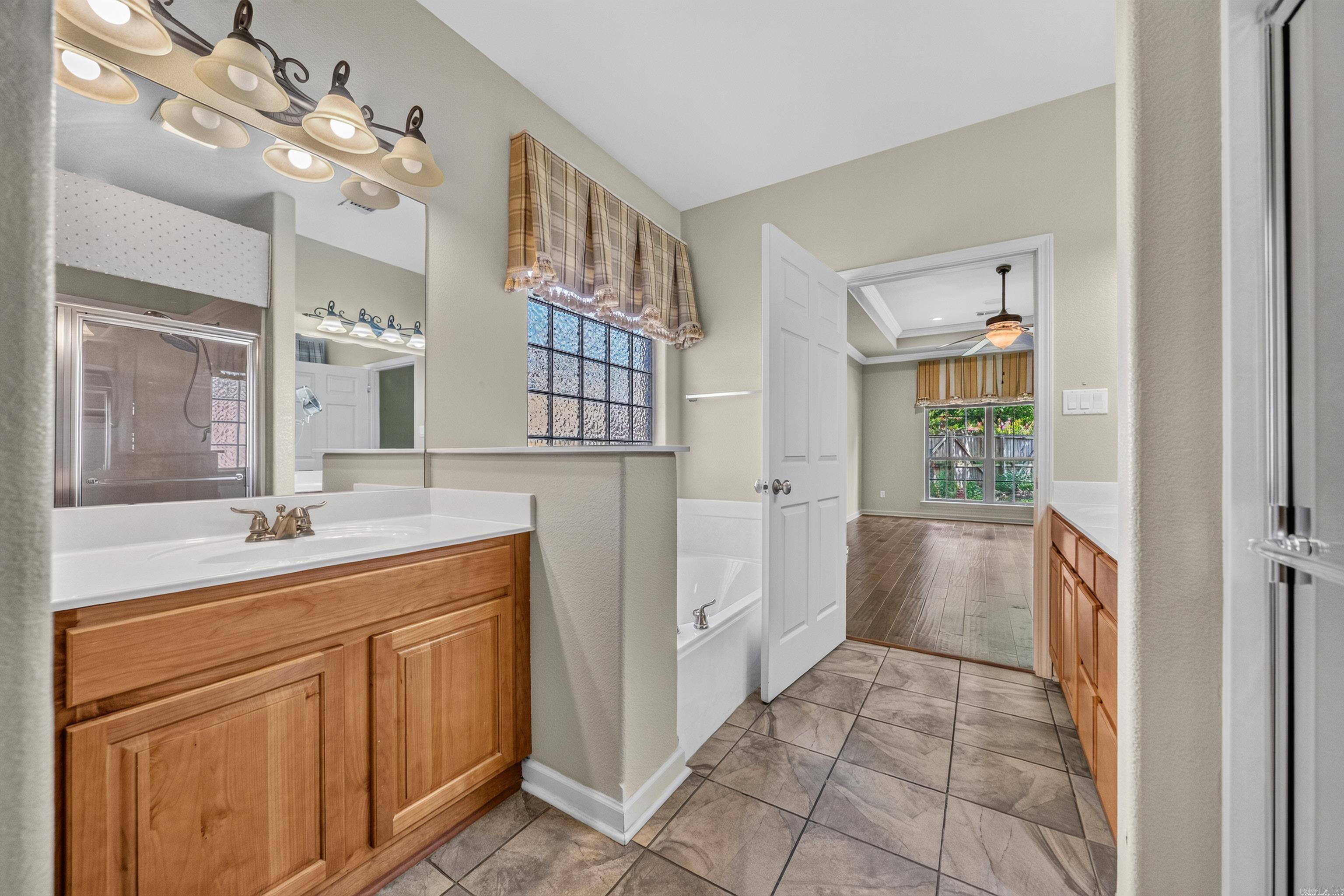 Bathroom with a wood vanity, tiled floor, tub, and glass shower; doorway opens to a room with a window.
