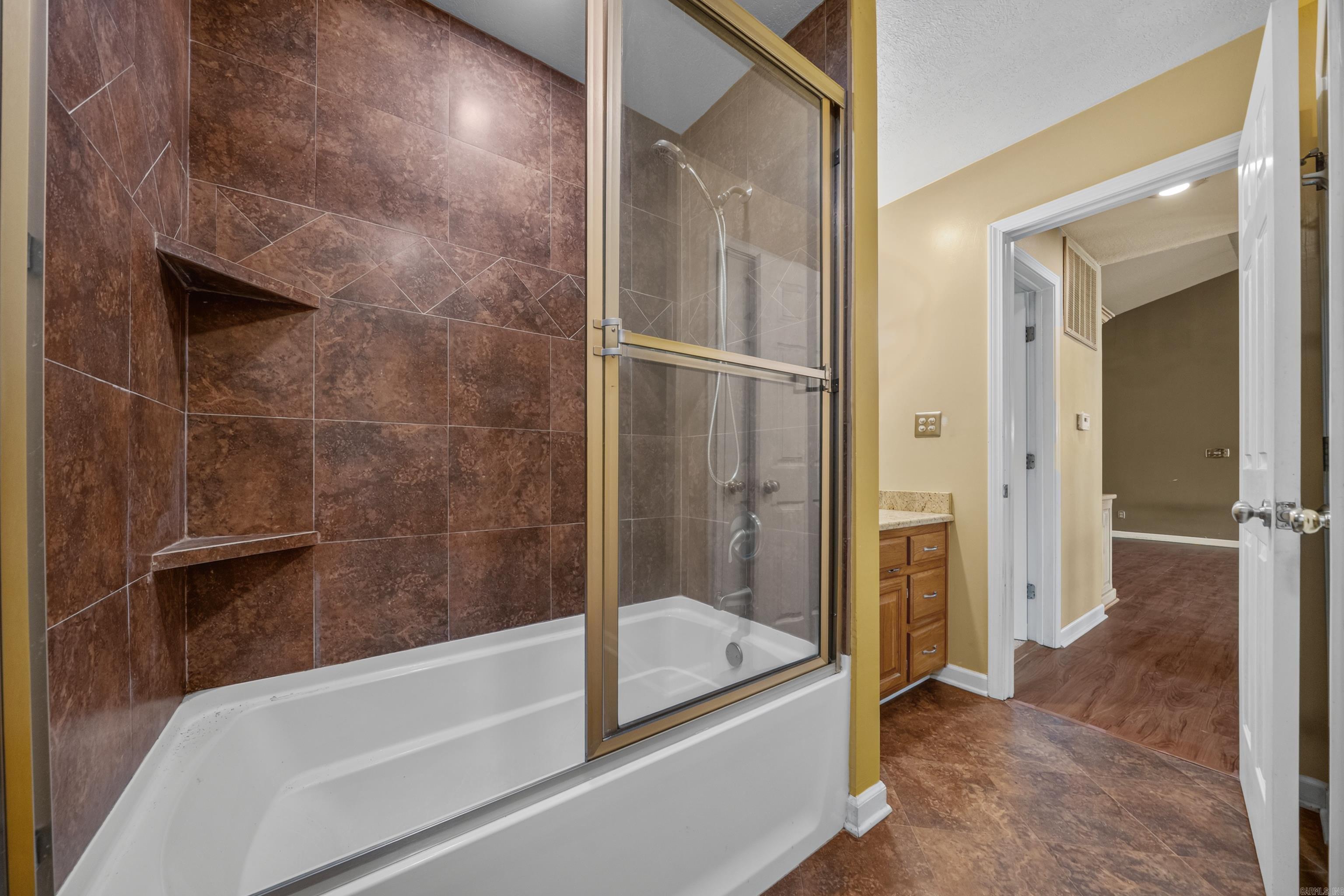 Bathroom with a glass-enclosed tub, brown tile, built-in shelves, and a wood vanity.