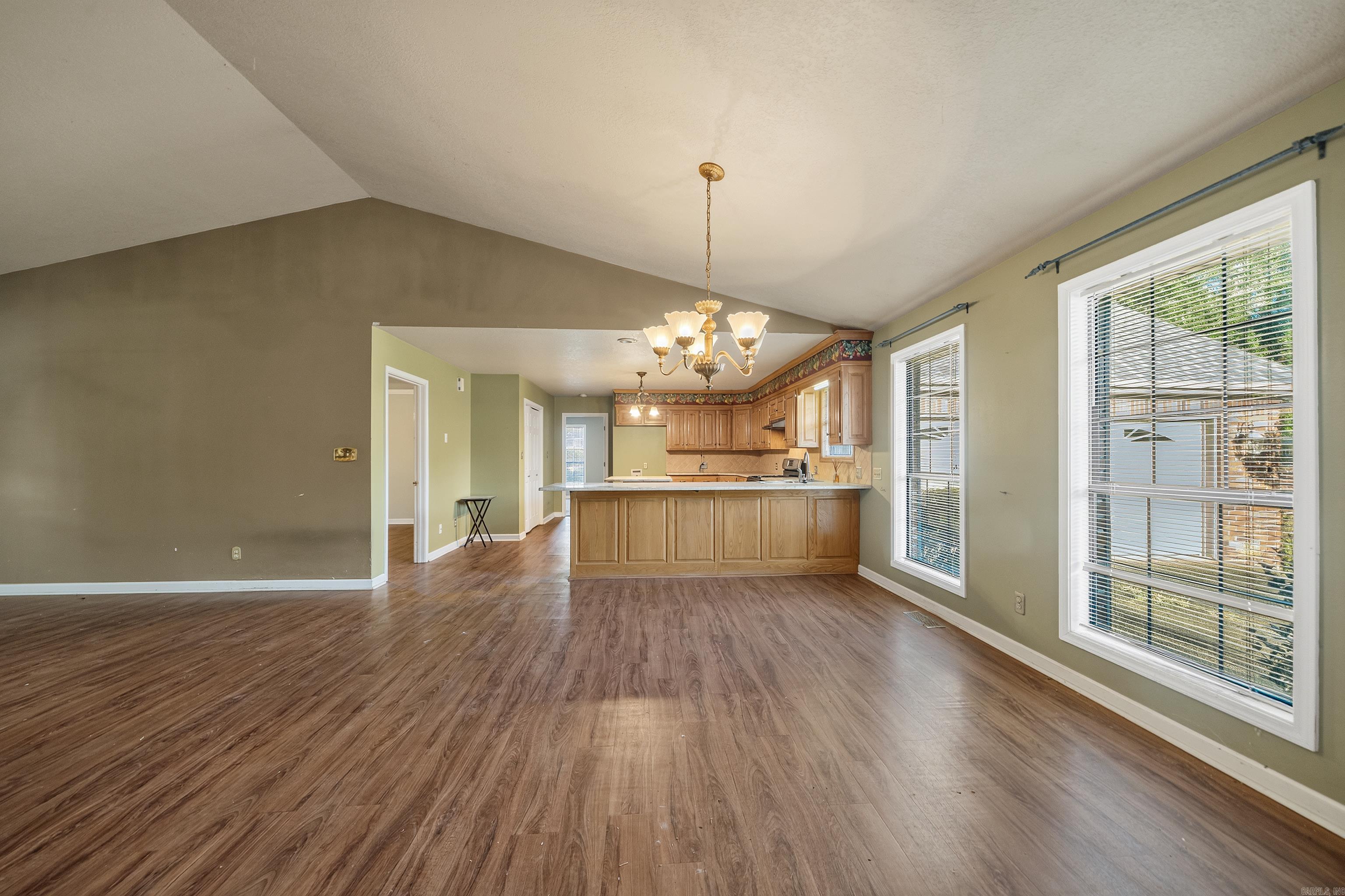 Vacant open-plan room with wood-look floors, vaulted ceiling, and a kitchen with light wood cabinets.