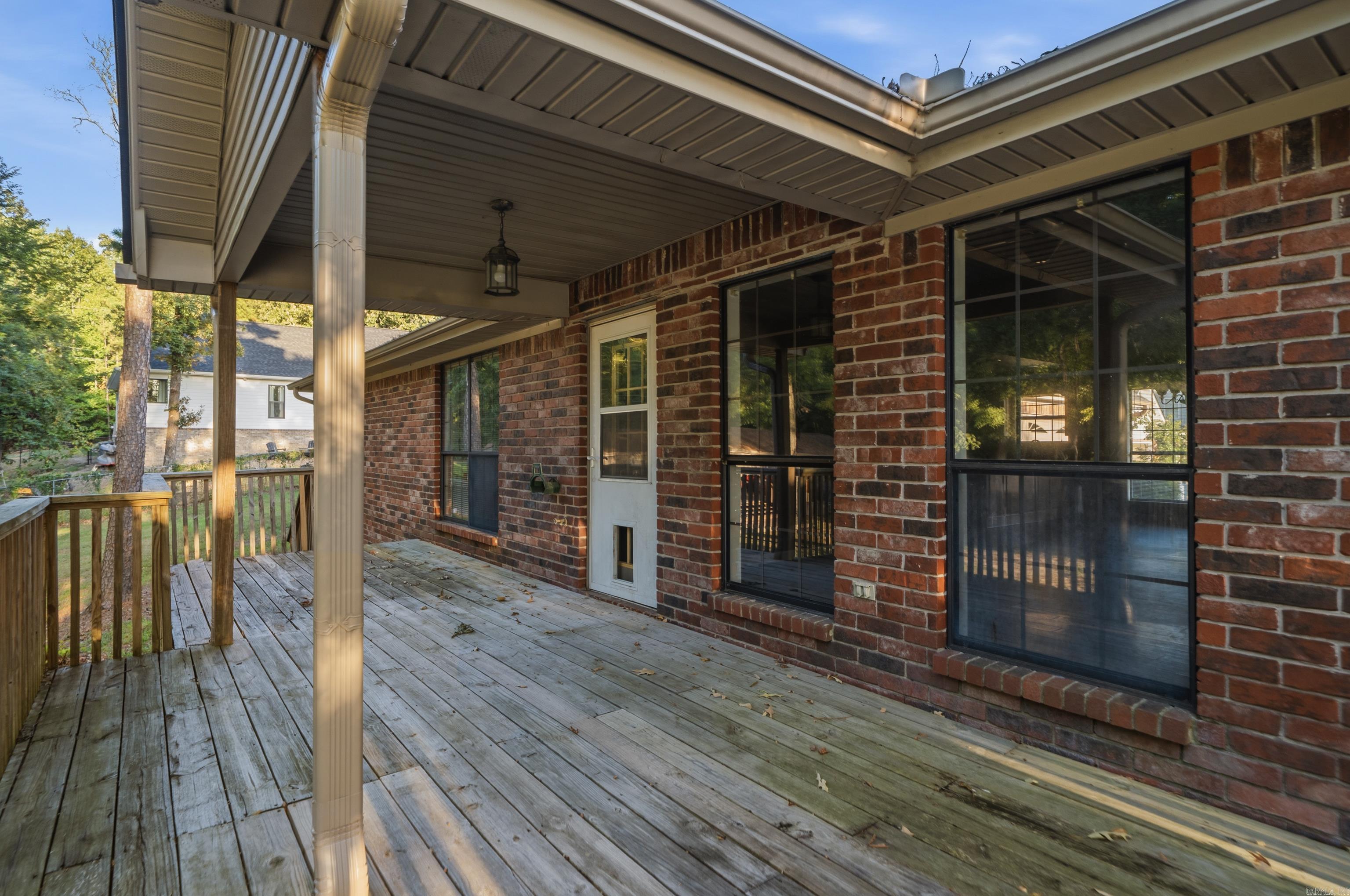 Covered wooden deck beside a brick house, with large windows and a wooded yard.