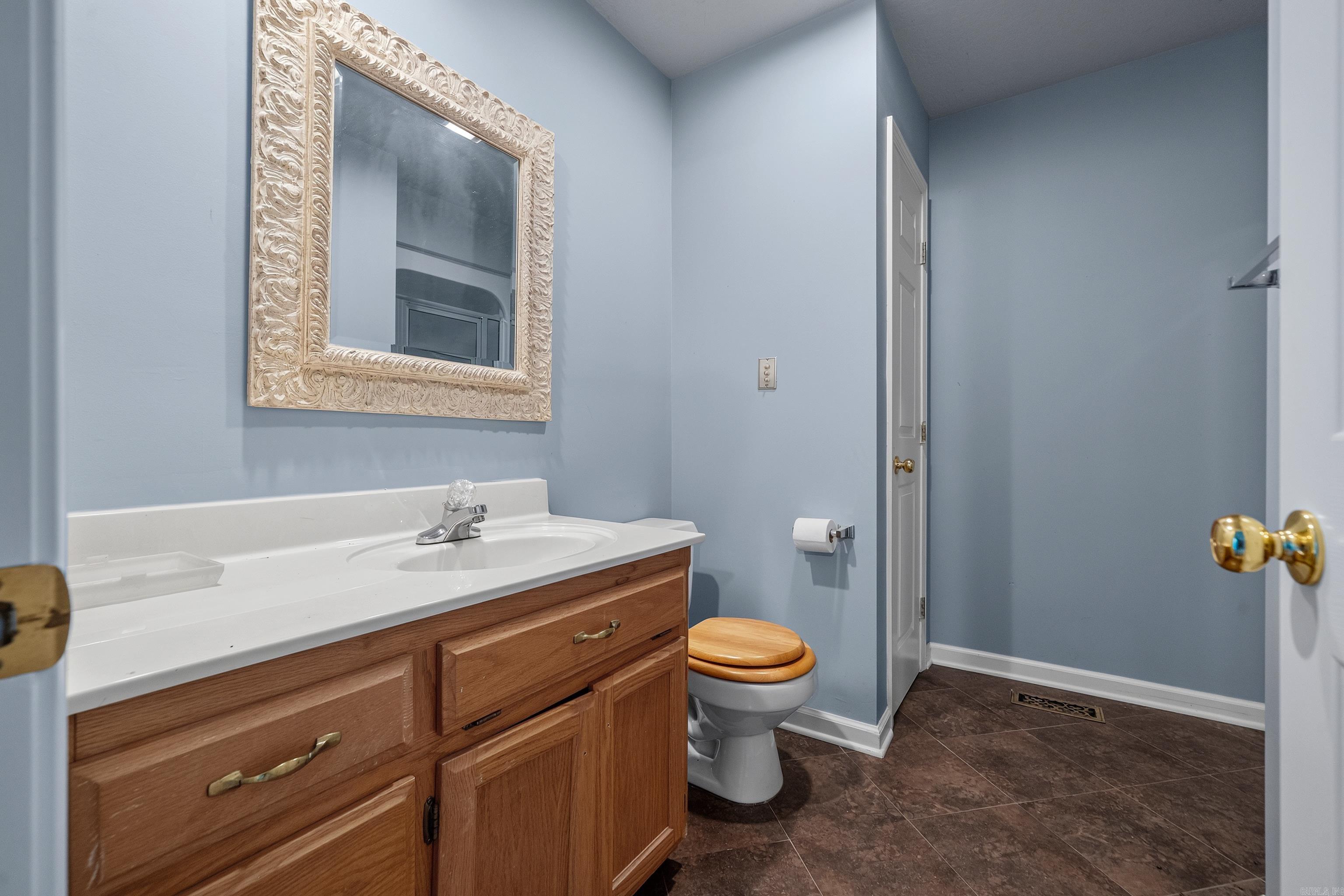 Bathroom with a wood vanity, ornate mirror, toilet with wooden seat, and dark tile floor.