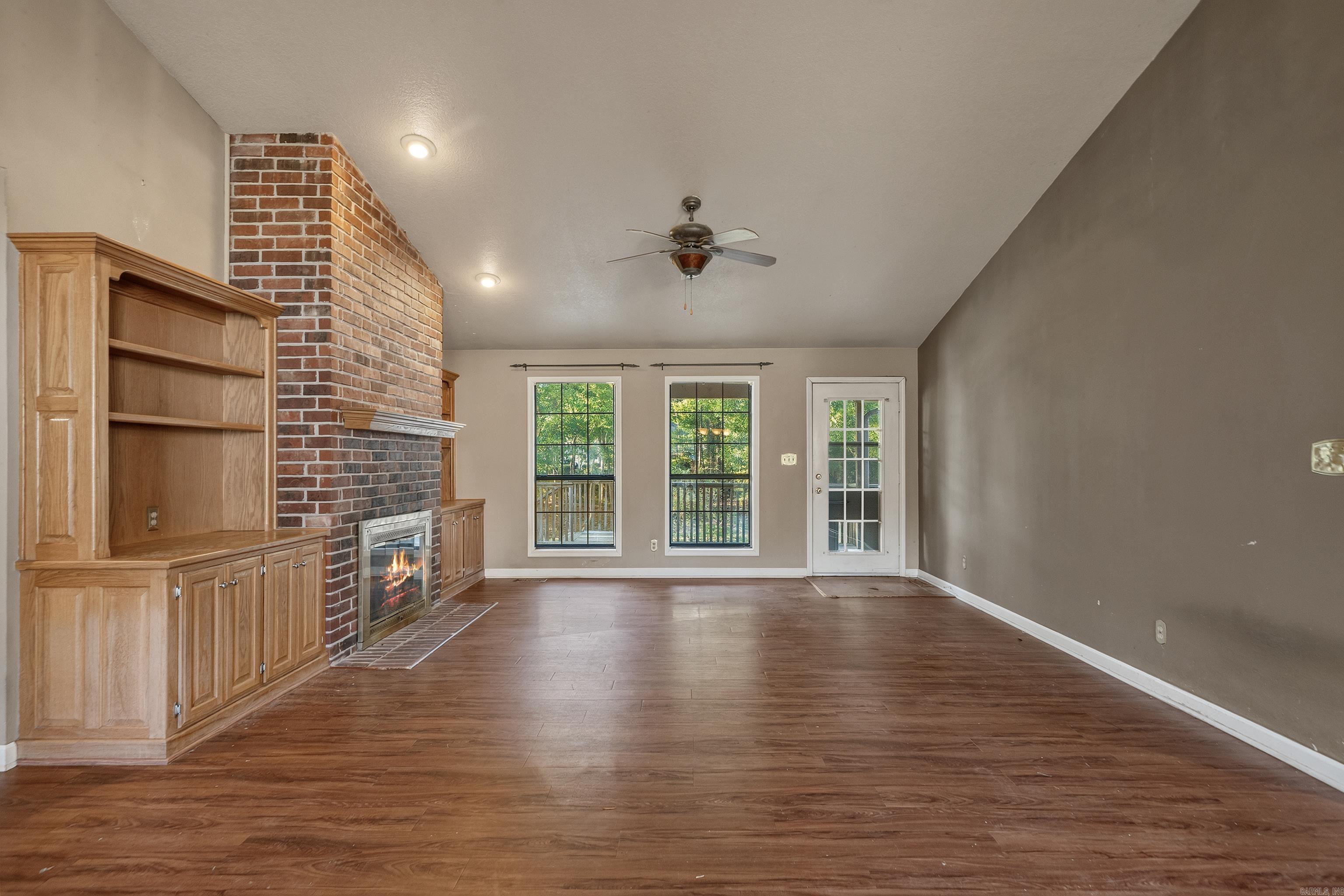 Empty living room with brick fireplace, built-in wood cabinetry, and windows overlooking trees.