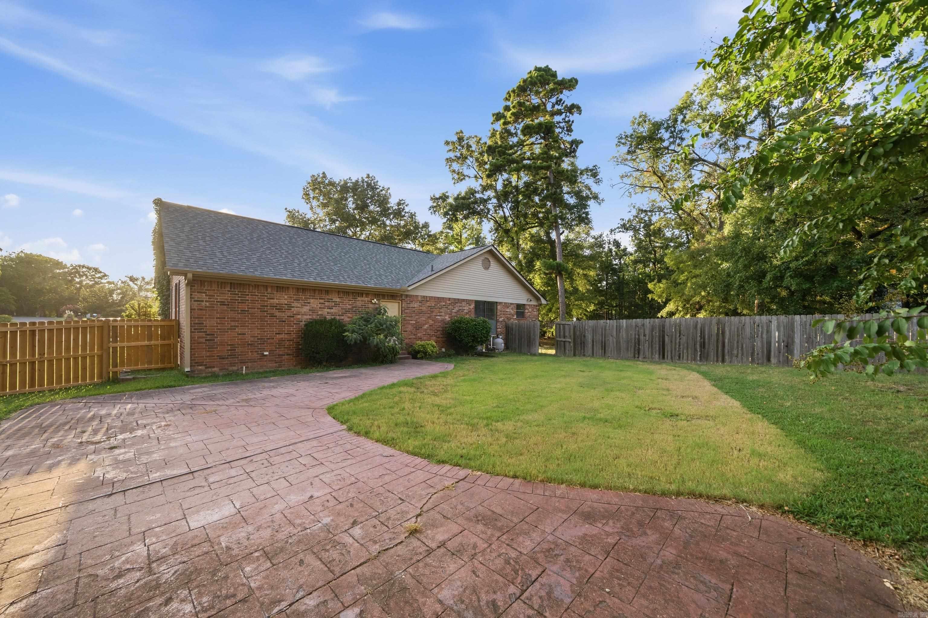 Brick home’s fenced backyard with a broad stamped-concrete patio, lawn, and mature trees.
