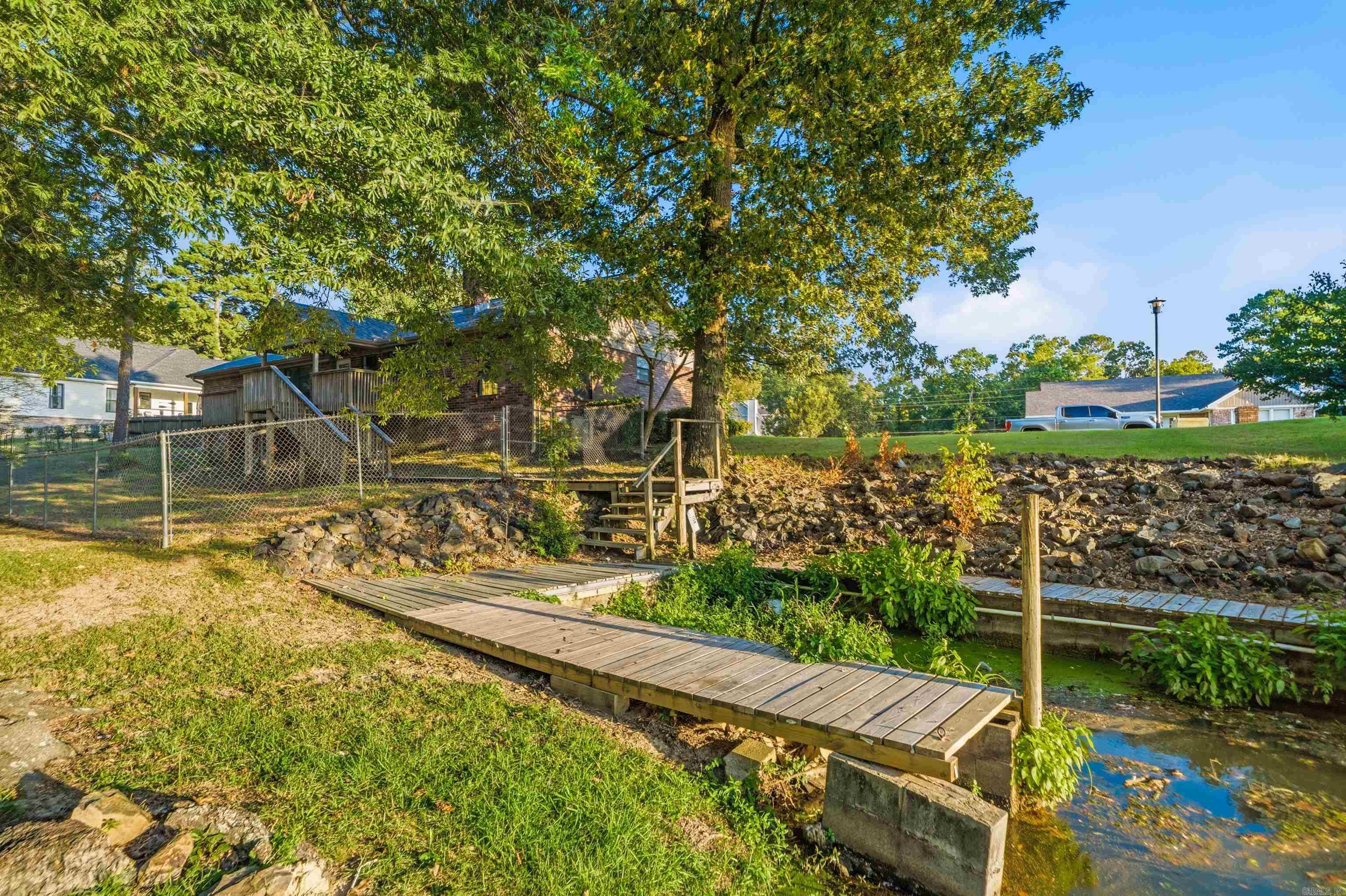 Brick house behind a fenced yard, with a wooden footbridge crossing a shallow creek.