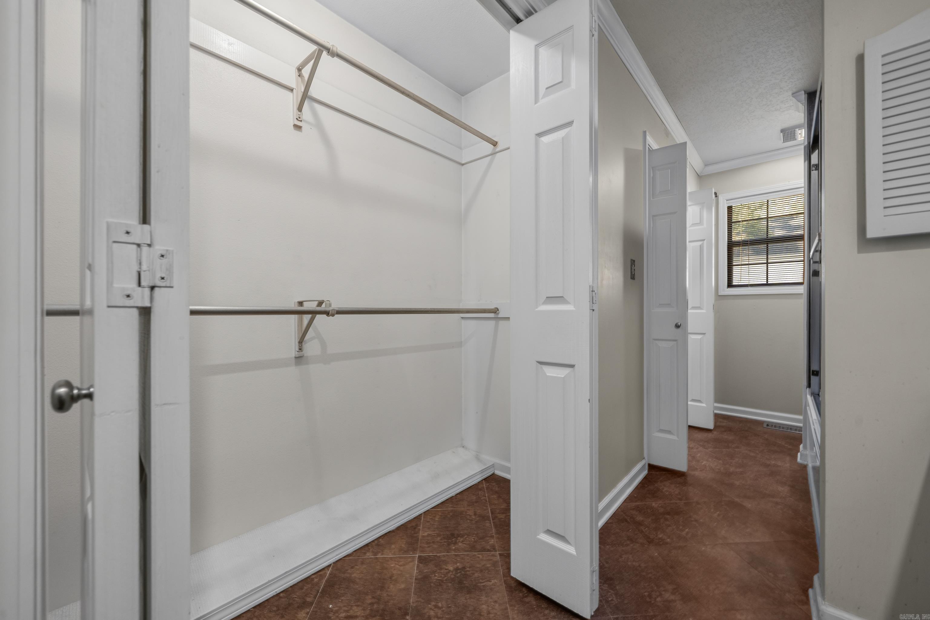 Empty walk-in closet with two metal hanging rods and a white platform beside a tiled hallway.