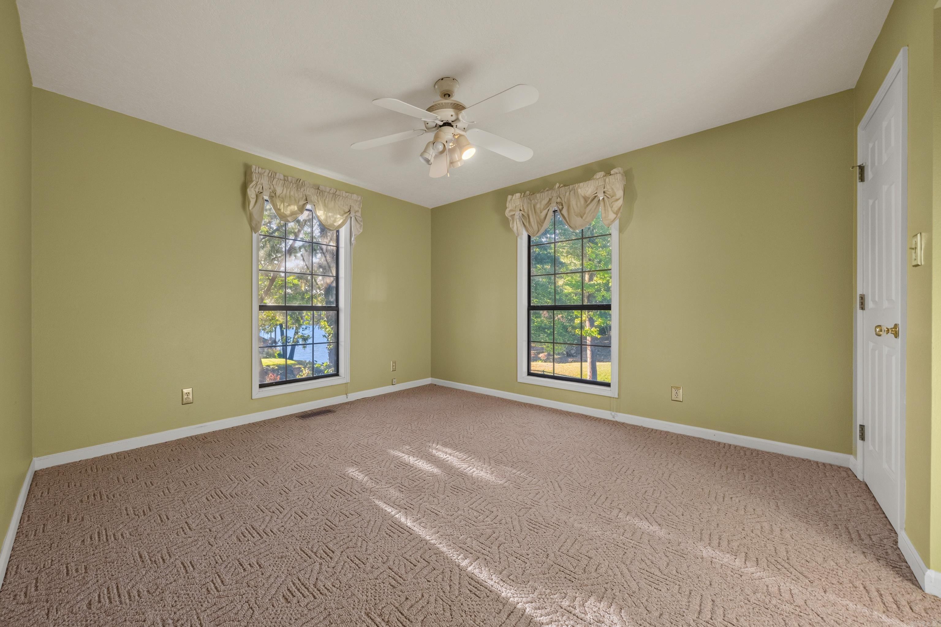 Empty bedroom with olive-green walls, patterned carpet, two curtained windows, and a ceiling fan.