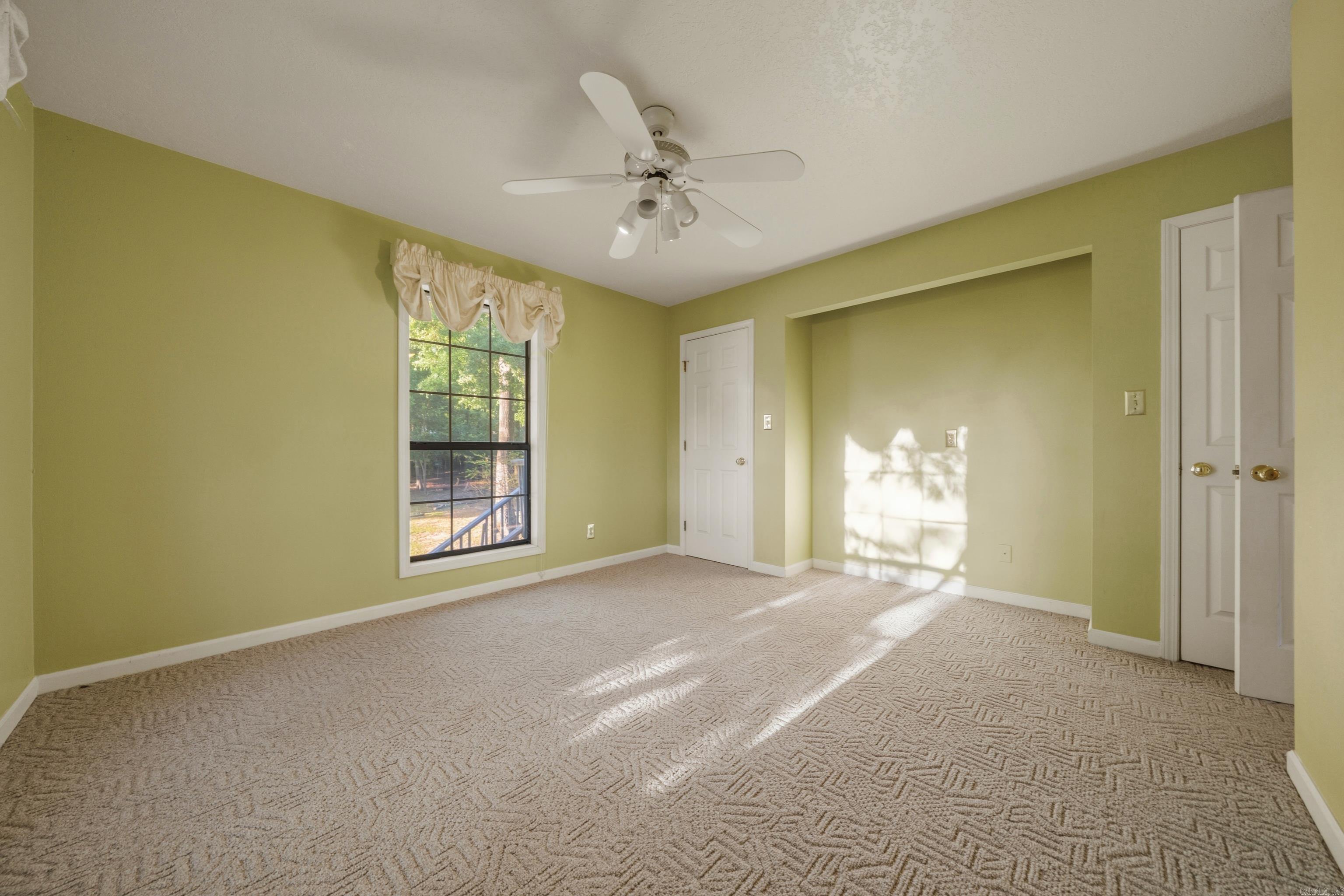 Empty bedroom with pale green walls, patterned carpet, and sunlight streaming through a large window.