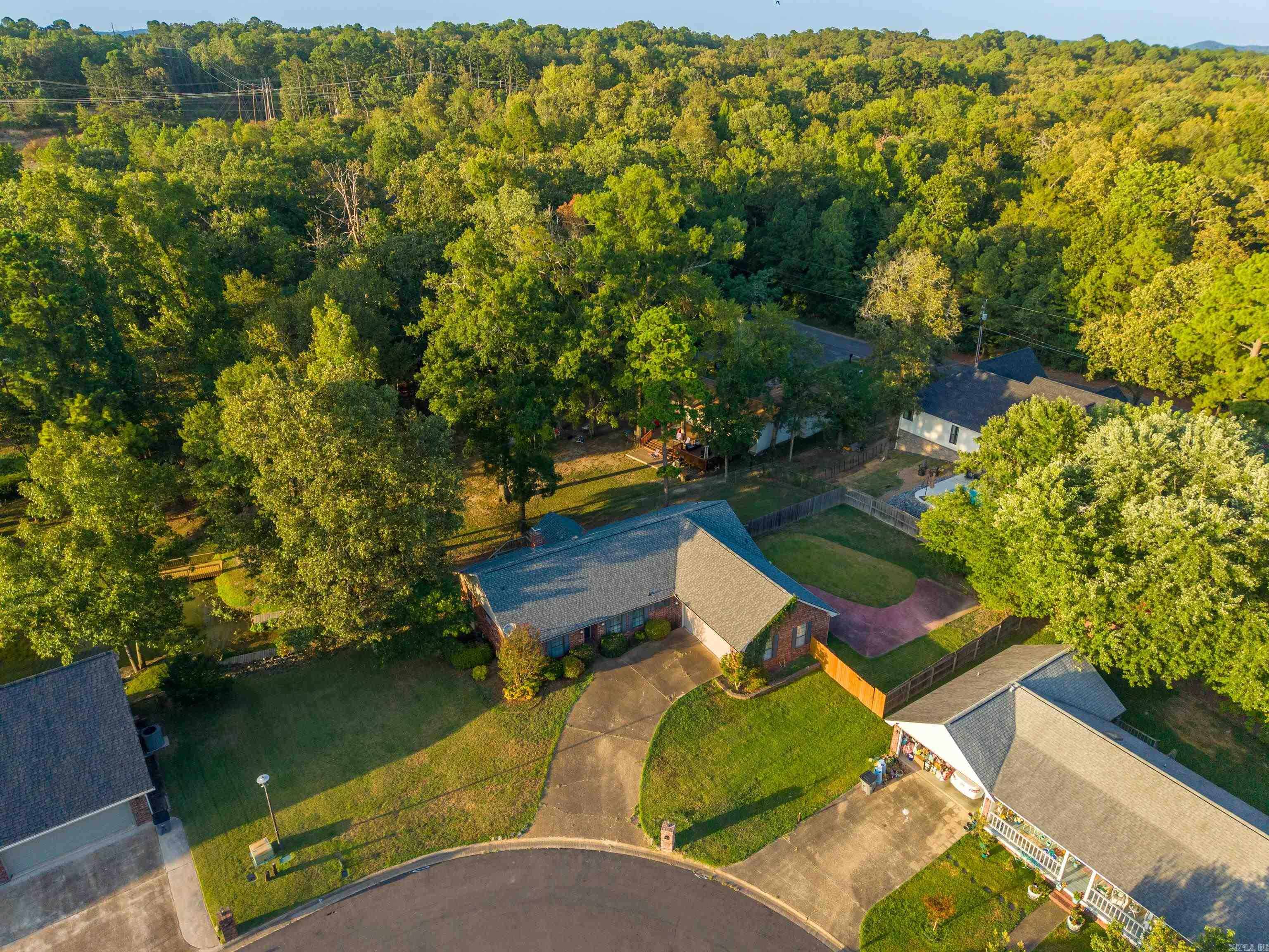 Aerial view of a brick home with a curved driveway and fenced yard beside dense woodland.