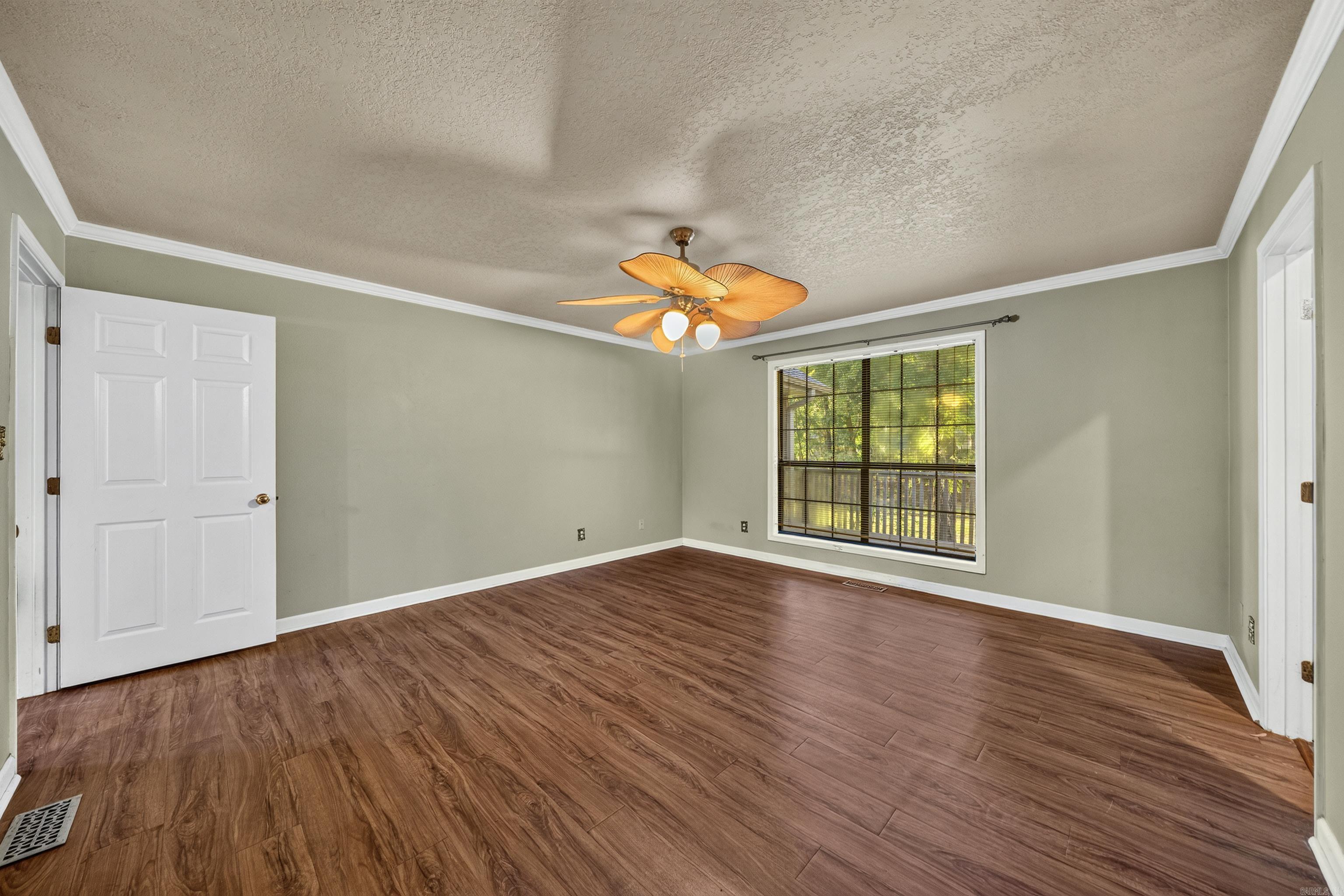 Empty room with wood-look flooring, a lit ceiling fan, and a window overlooking trees.
