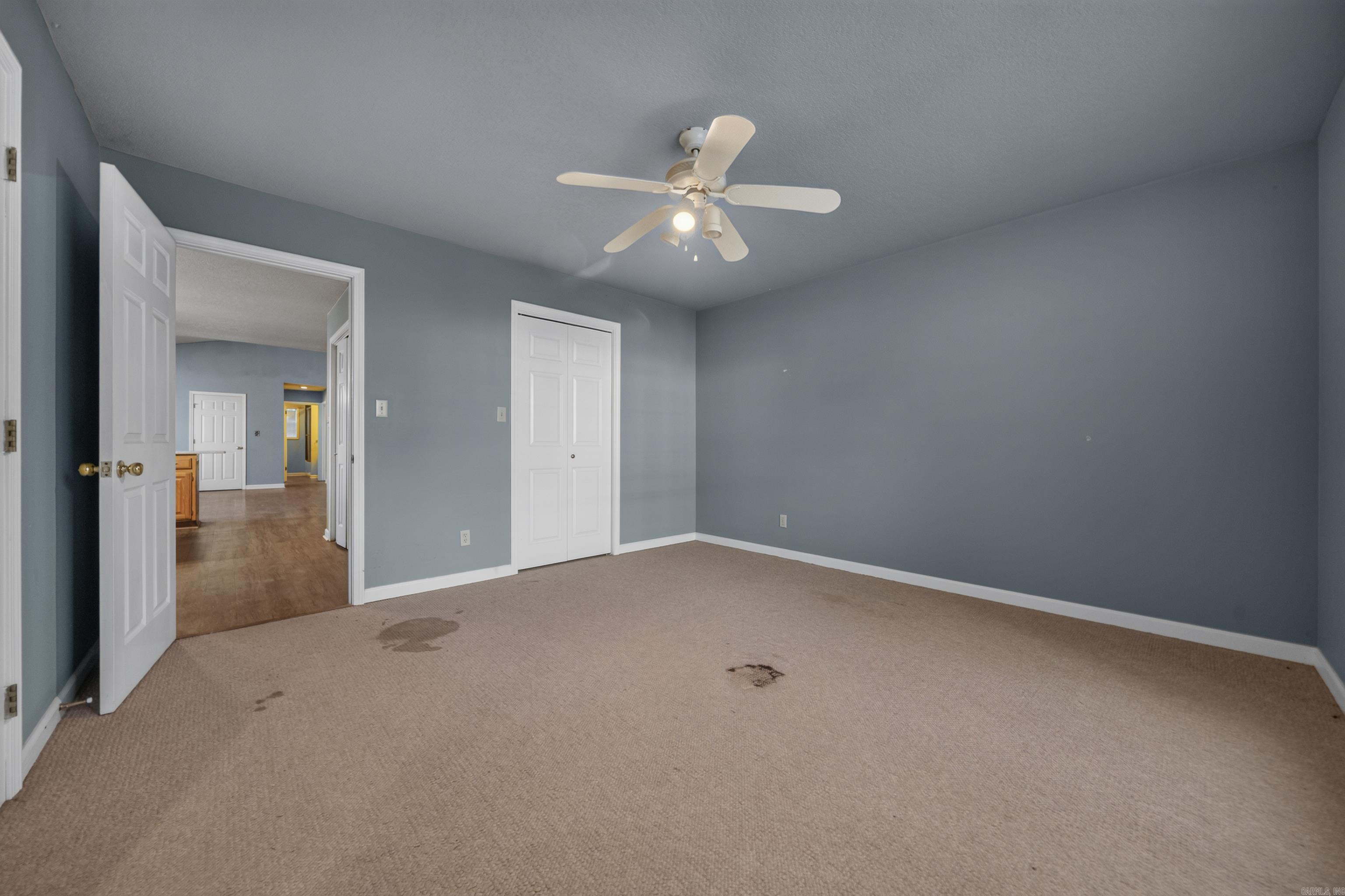 Empty bedroom with blue-gray walls, stained beige carpet, white closet doors, and a ceiling fan.
