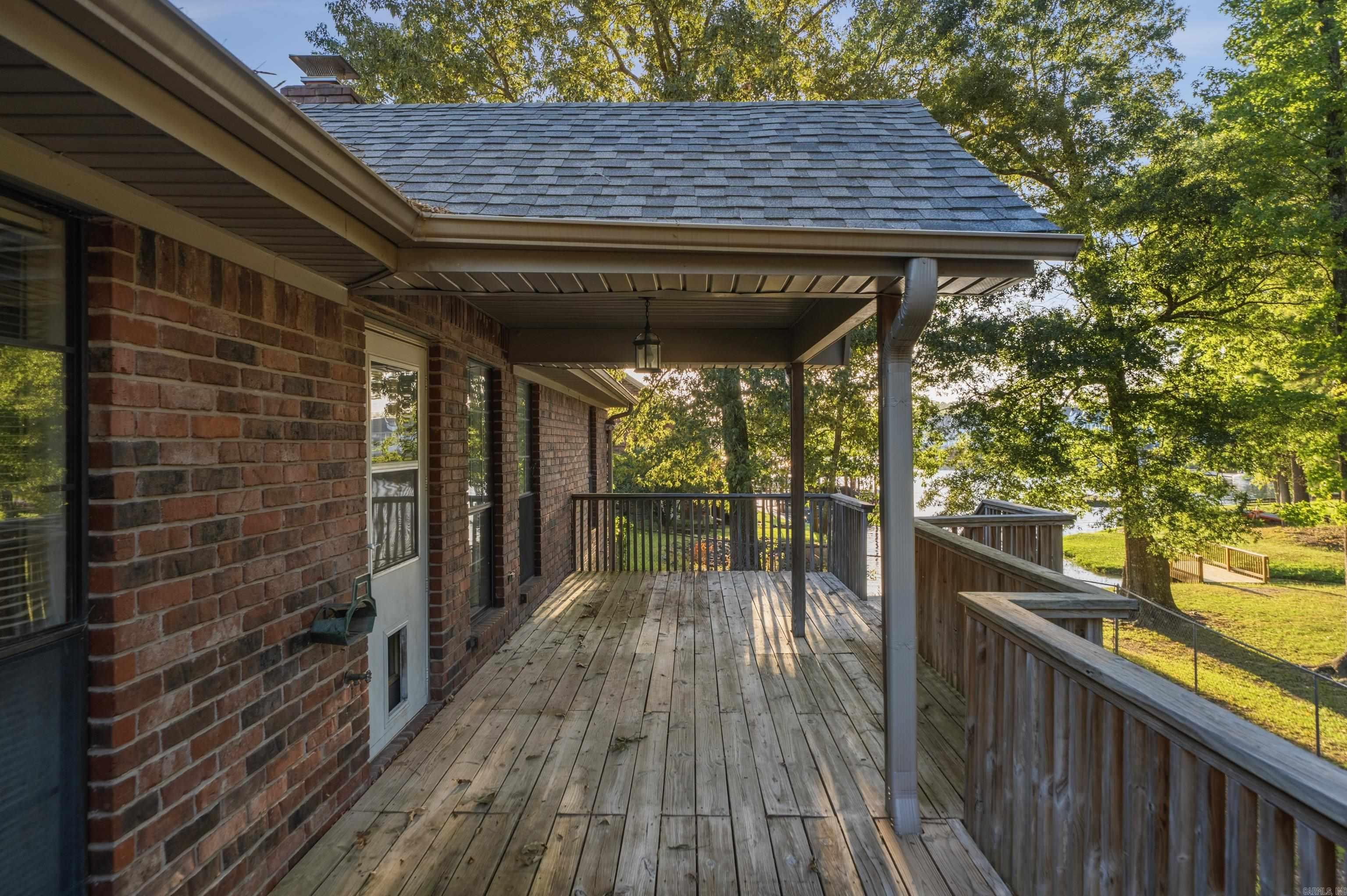 Covered wooden deck beside a brick house, with trees and water beyond the railing.