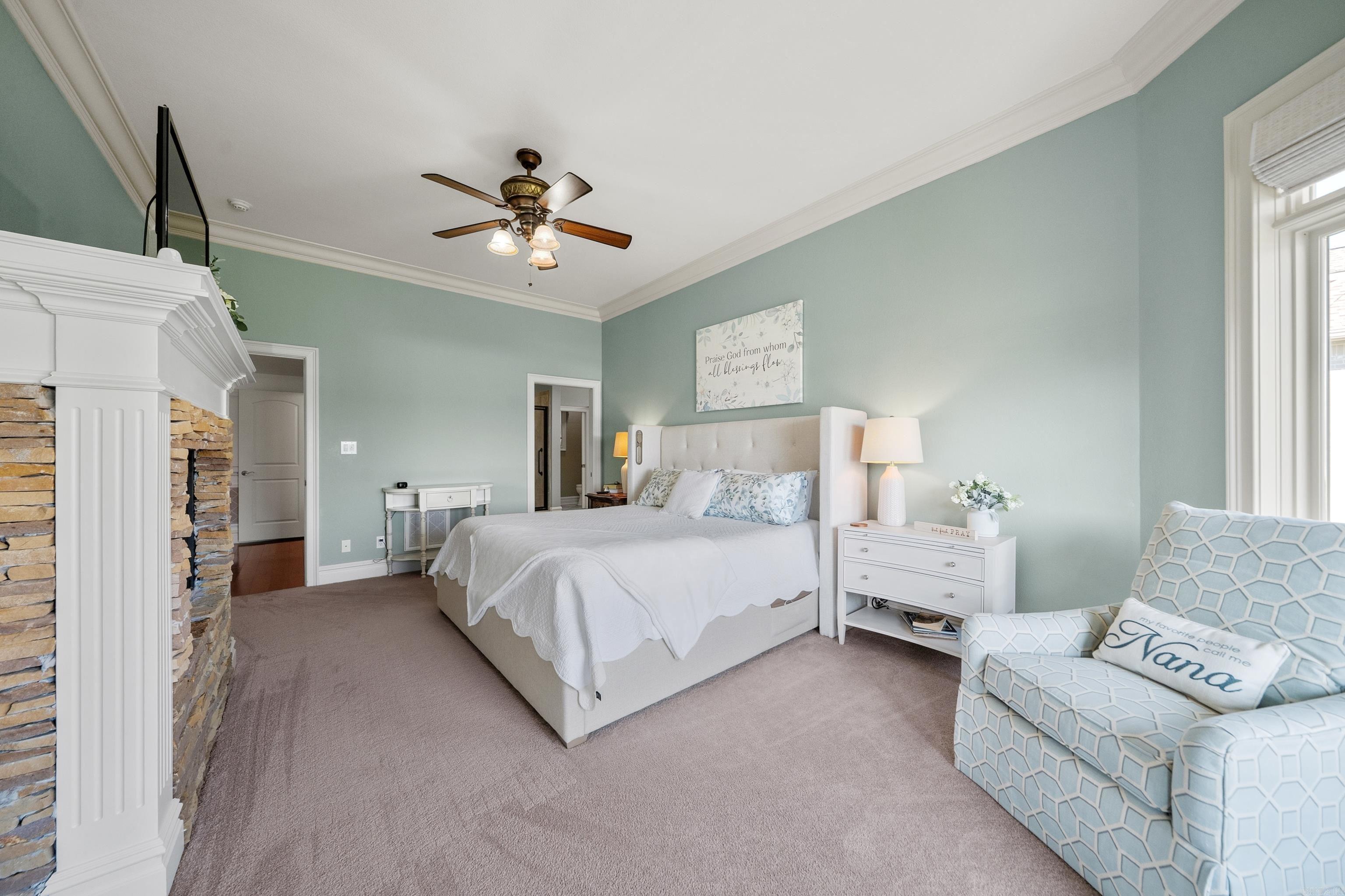 Bedroom with a white bed, stone fireplace, sage walls, and patterned armchair by a bright window.