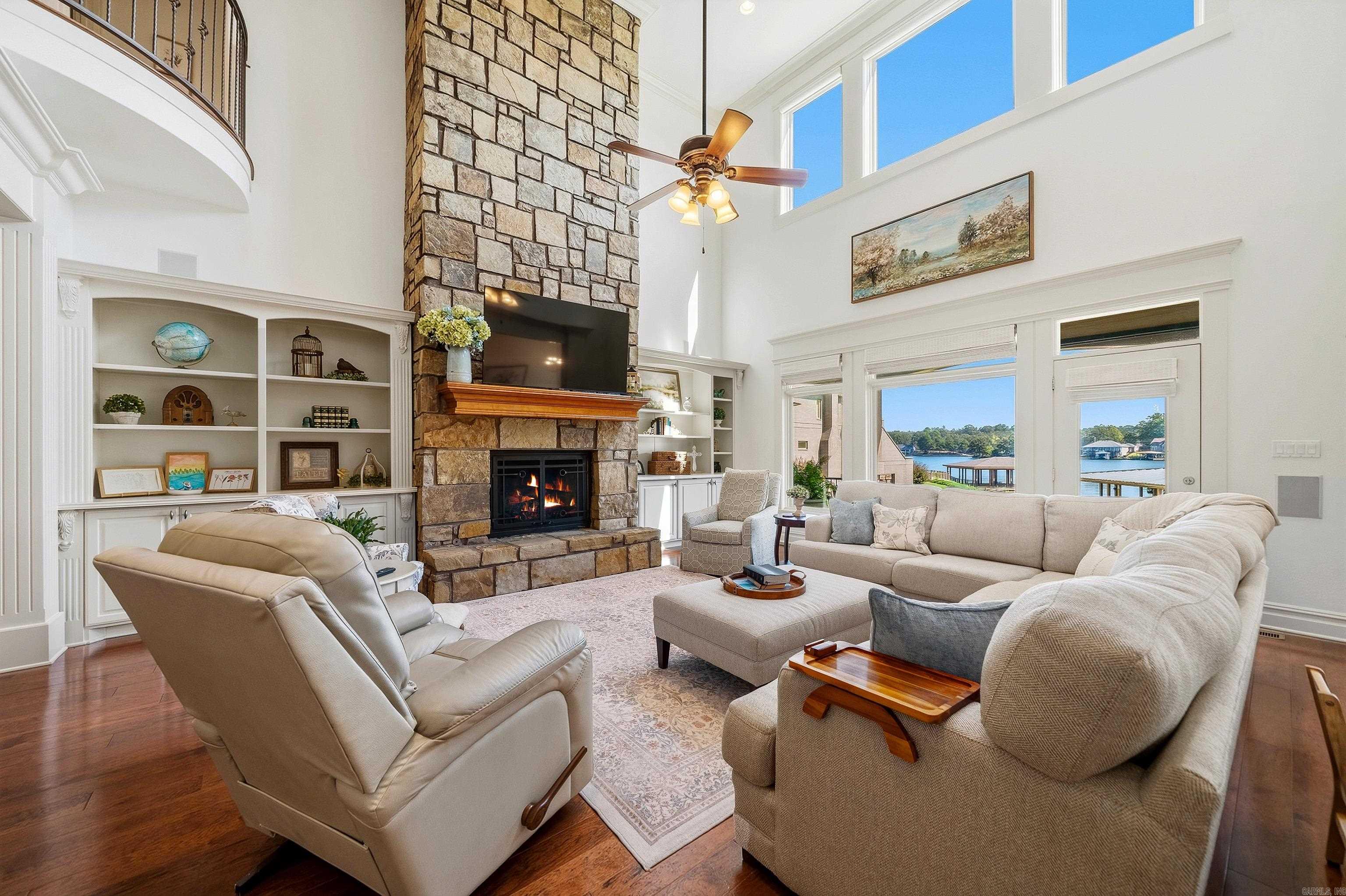 Living room with a tall stone fireplace, double-height windows, and a view of the lake.