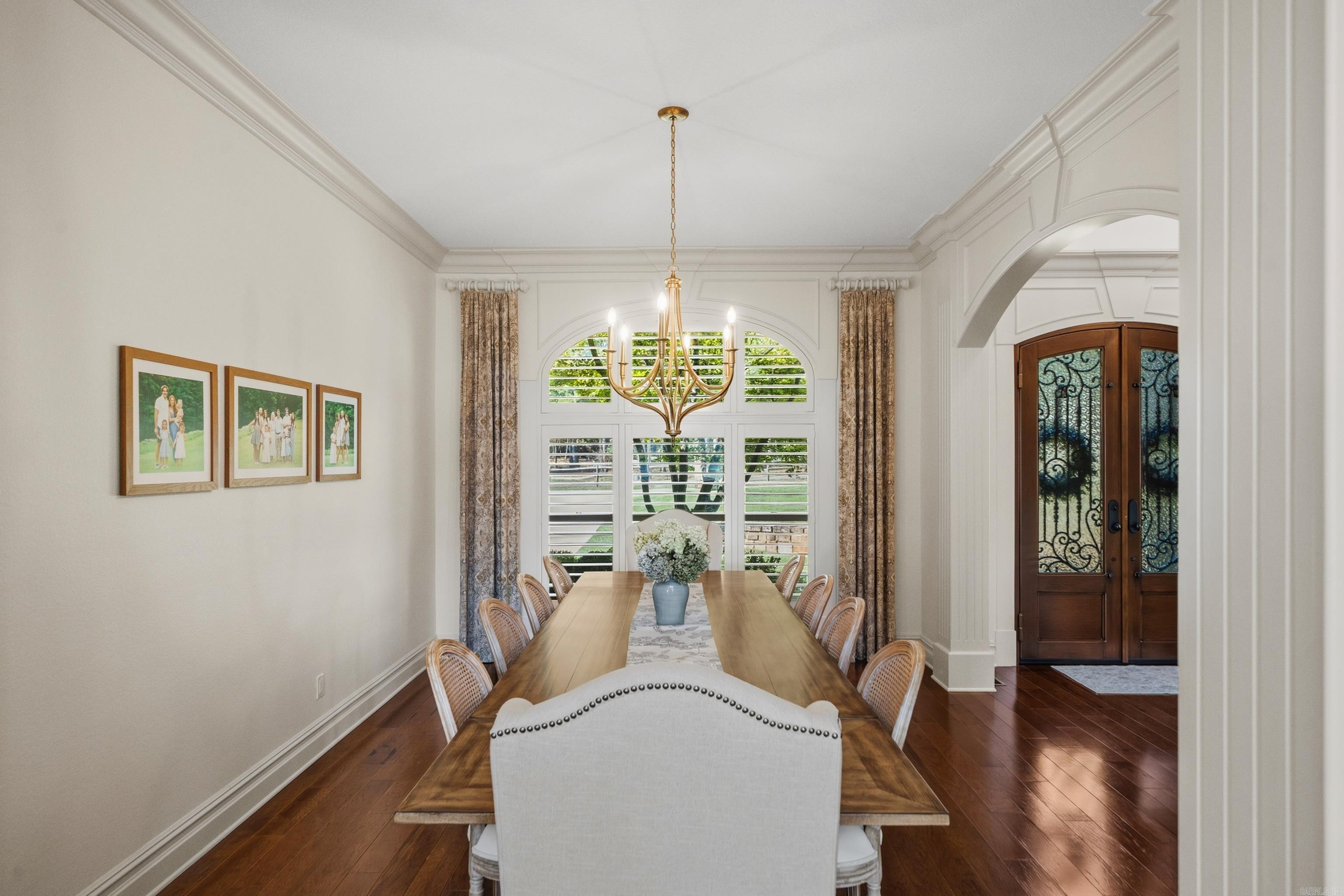 Dining room with a long wooden table, brass chandelier, arched windows, and ornate double doors.