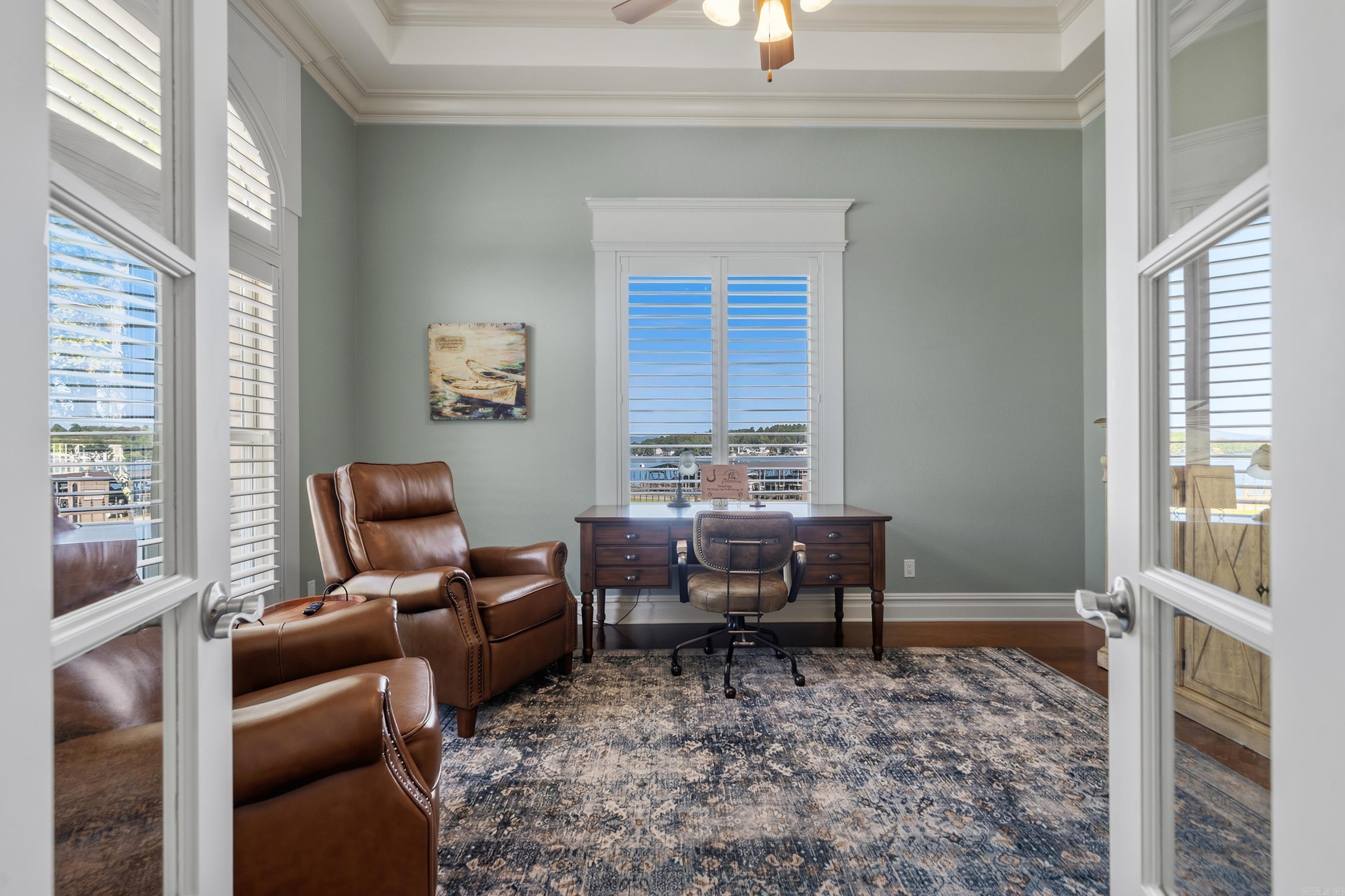 Furnished home office with a dark wood desk, leather armchairs, and windows overlooking the water.