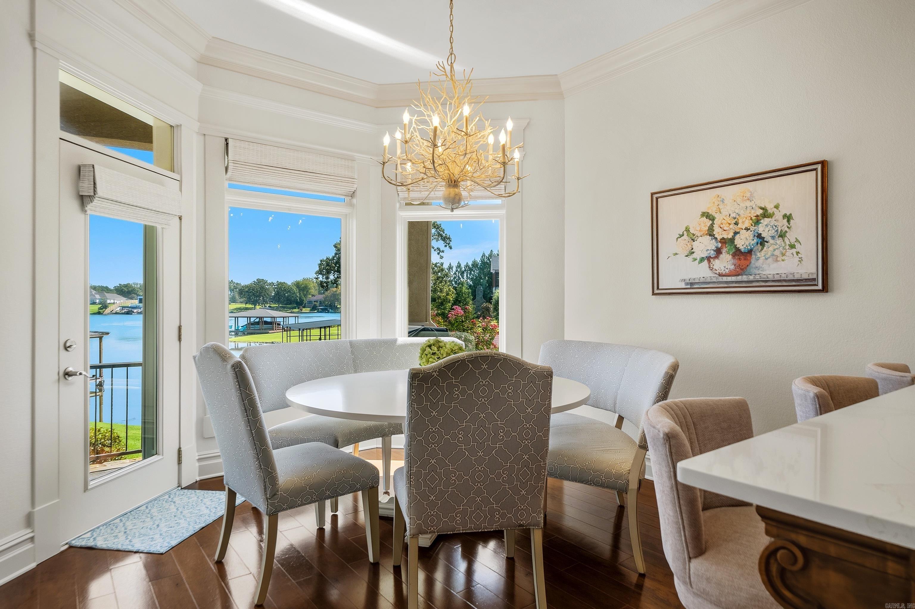 Dining area with a round table and chandelier, overlooking the water through large windows.