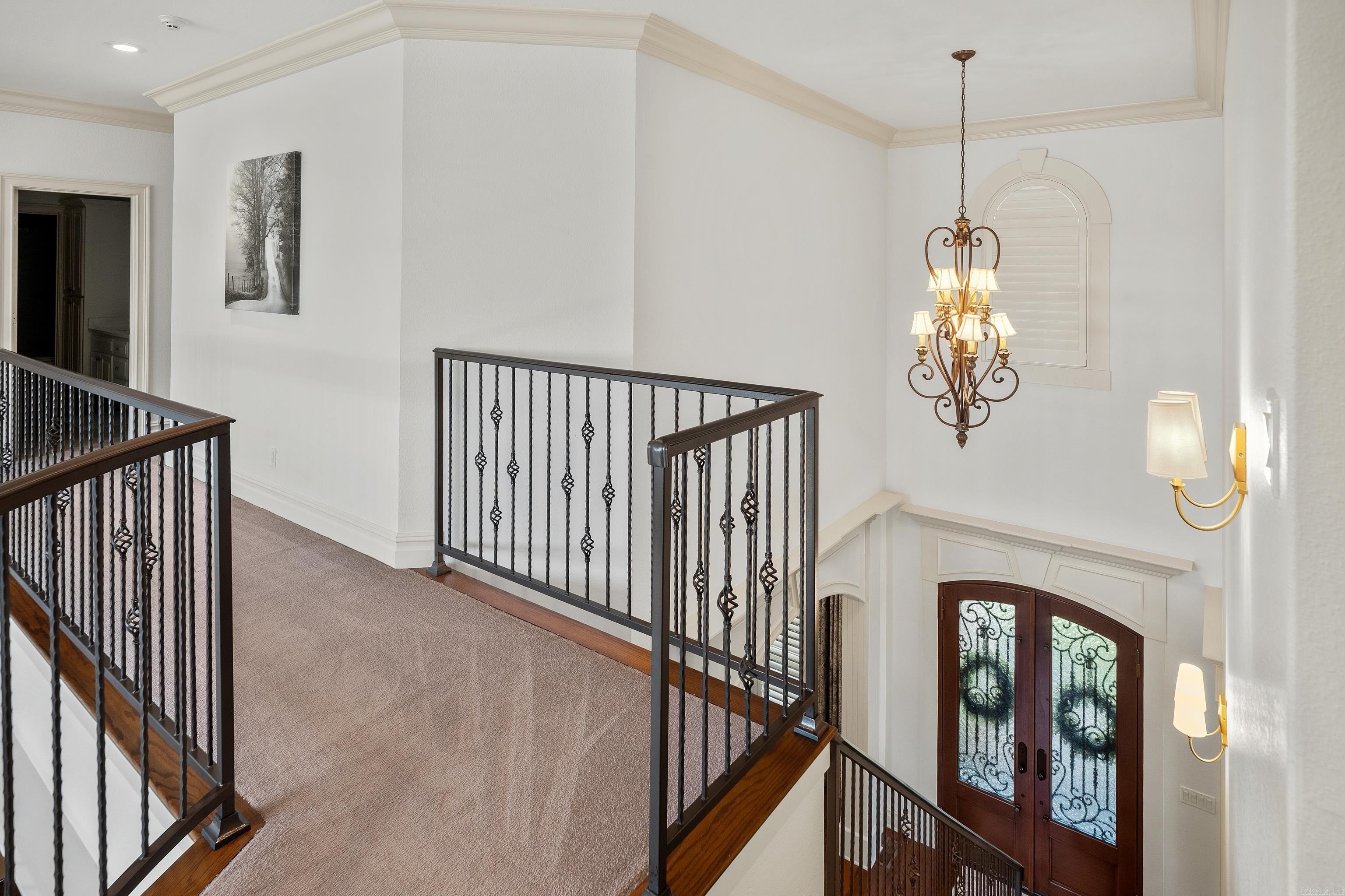 Upstairs landing overlooking a foyer, with ornate iron railings, a chandelier, and decorative glass entry doors.