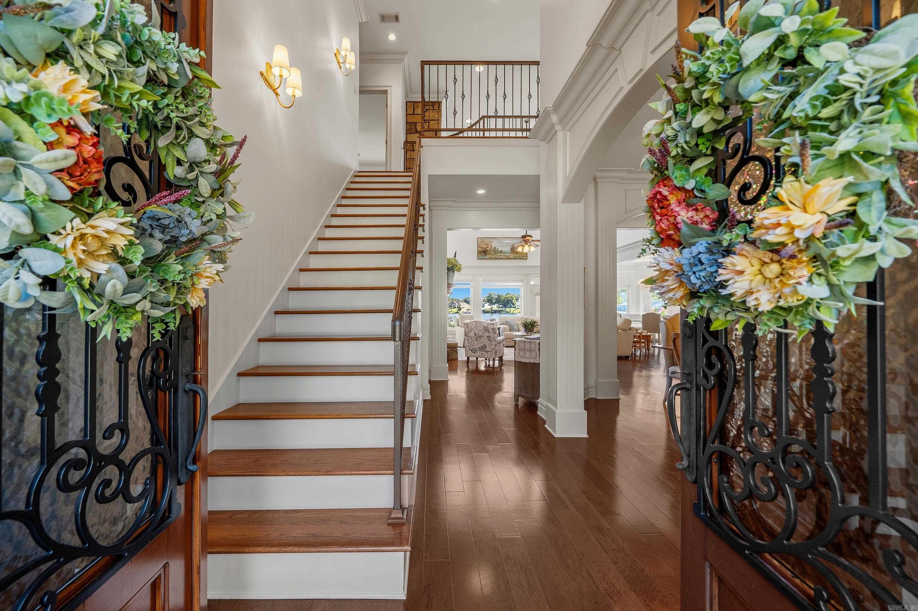 Foyer with a wood-and-white staircase, dark floors, and floral arrangements framing ornate entry doors.